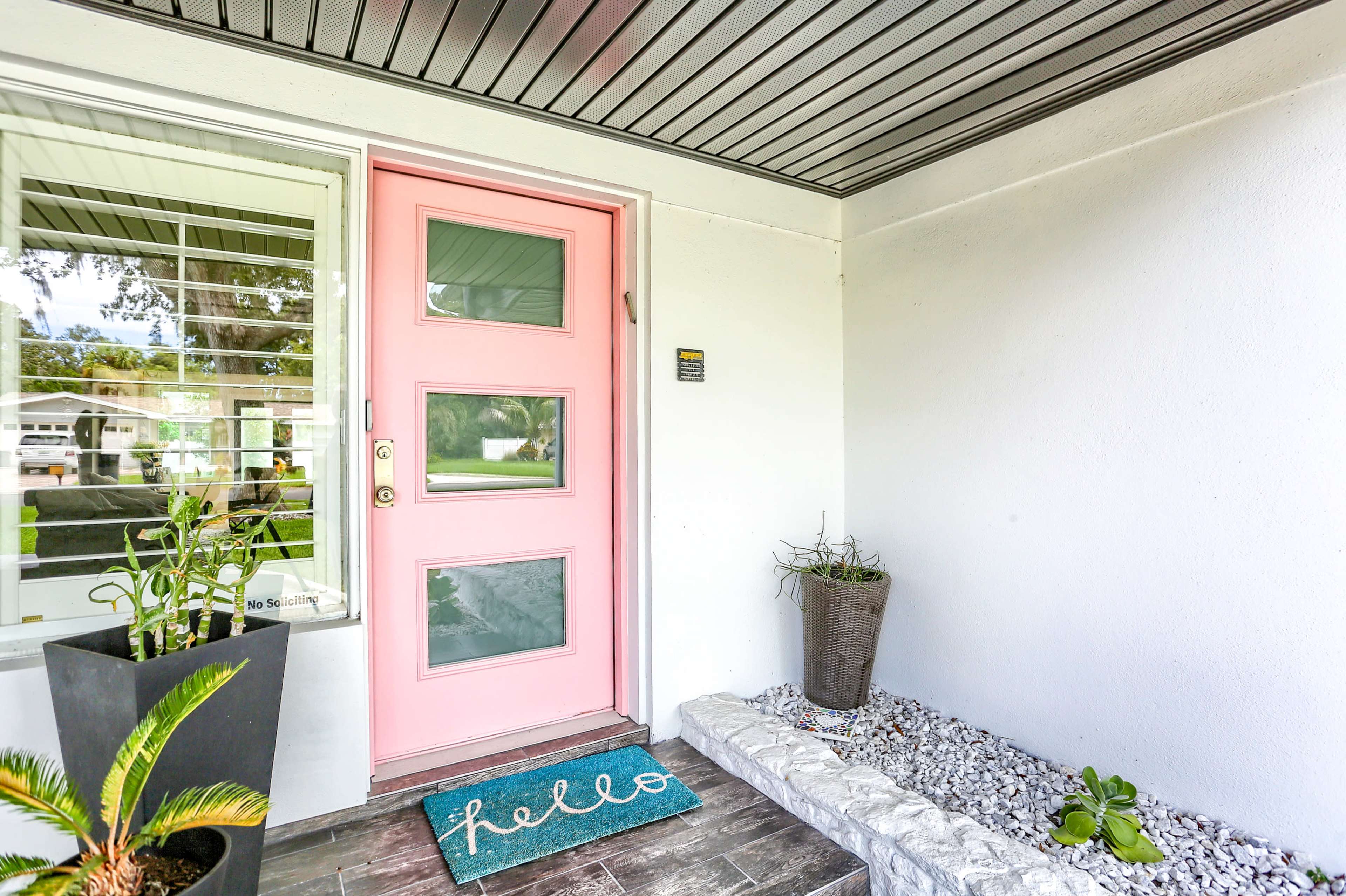 A modern house entrance featuring a pink front door, a welcome mat with "hello," potted plants, and a stone-lined planter.
