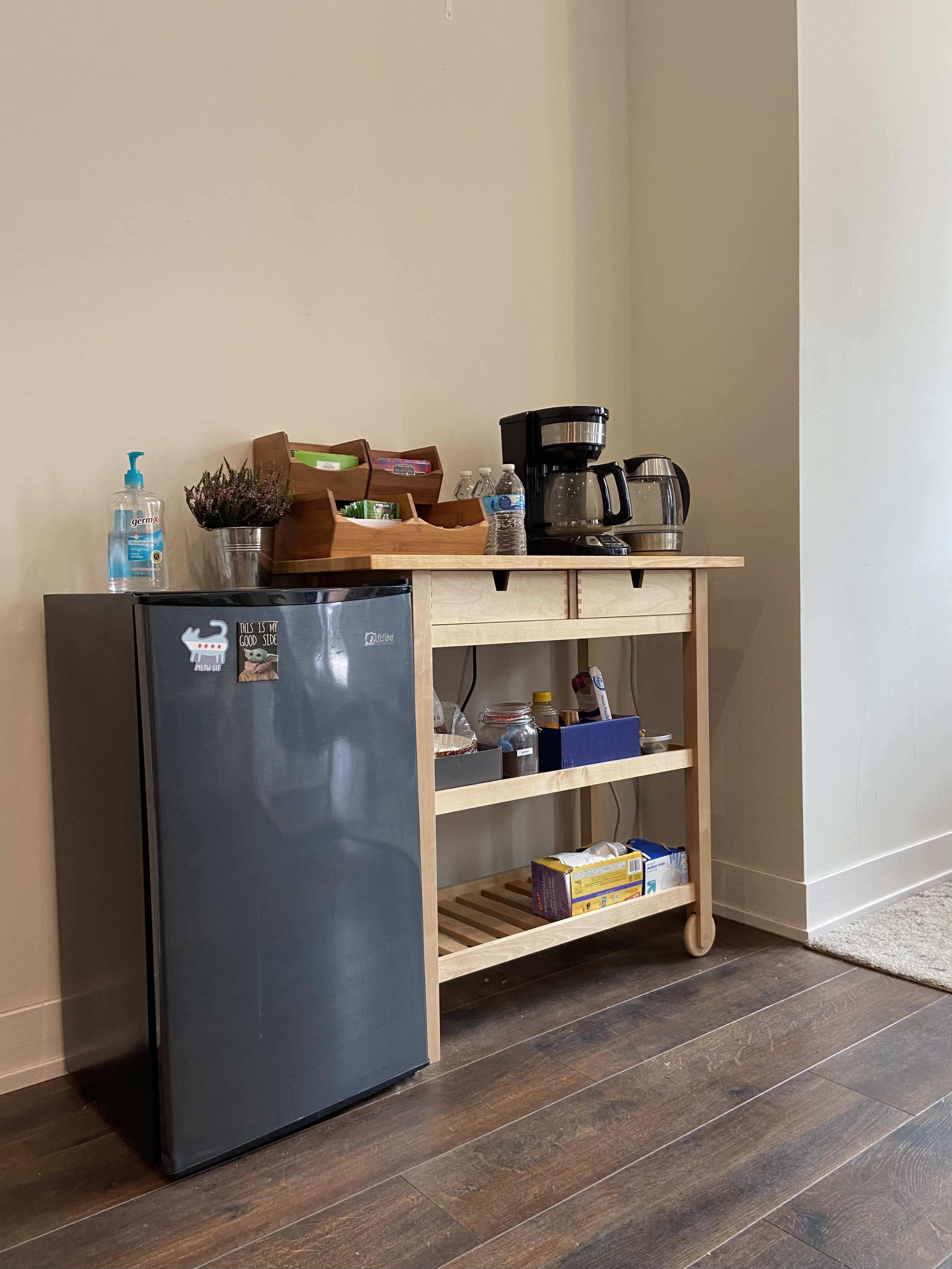A small kitchen area featuring a black refrigerator, a wooden shelf with a coffee maker, and various food items.