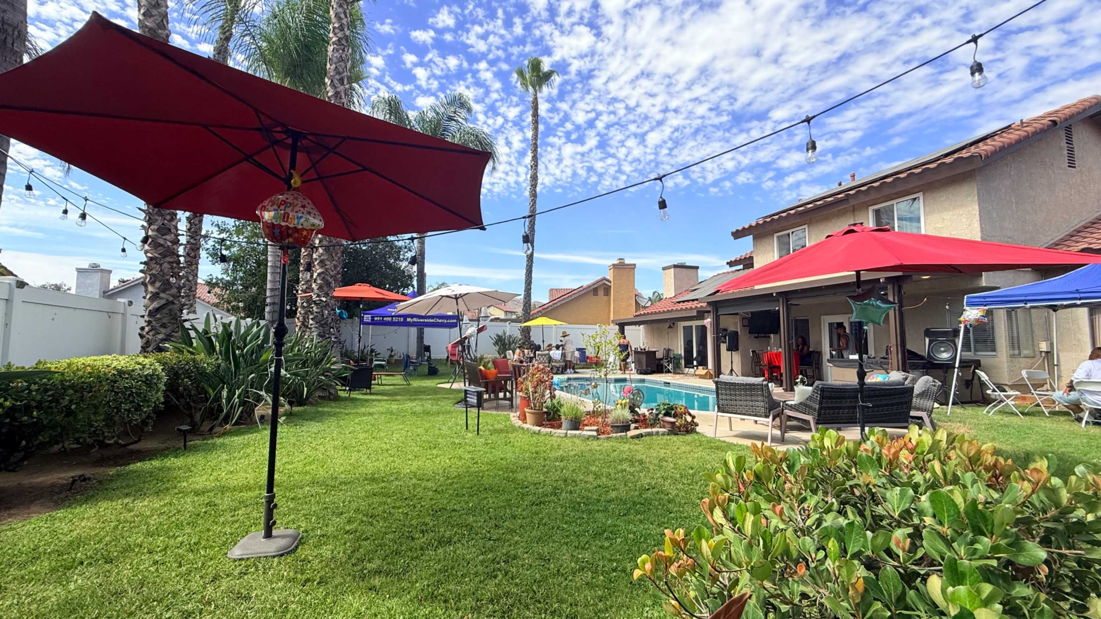 The image shows a vibrant backyard with a swimming pool, several umbrellas, and various outdoor seating areas surrounded by lush green grass and palm trees.