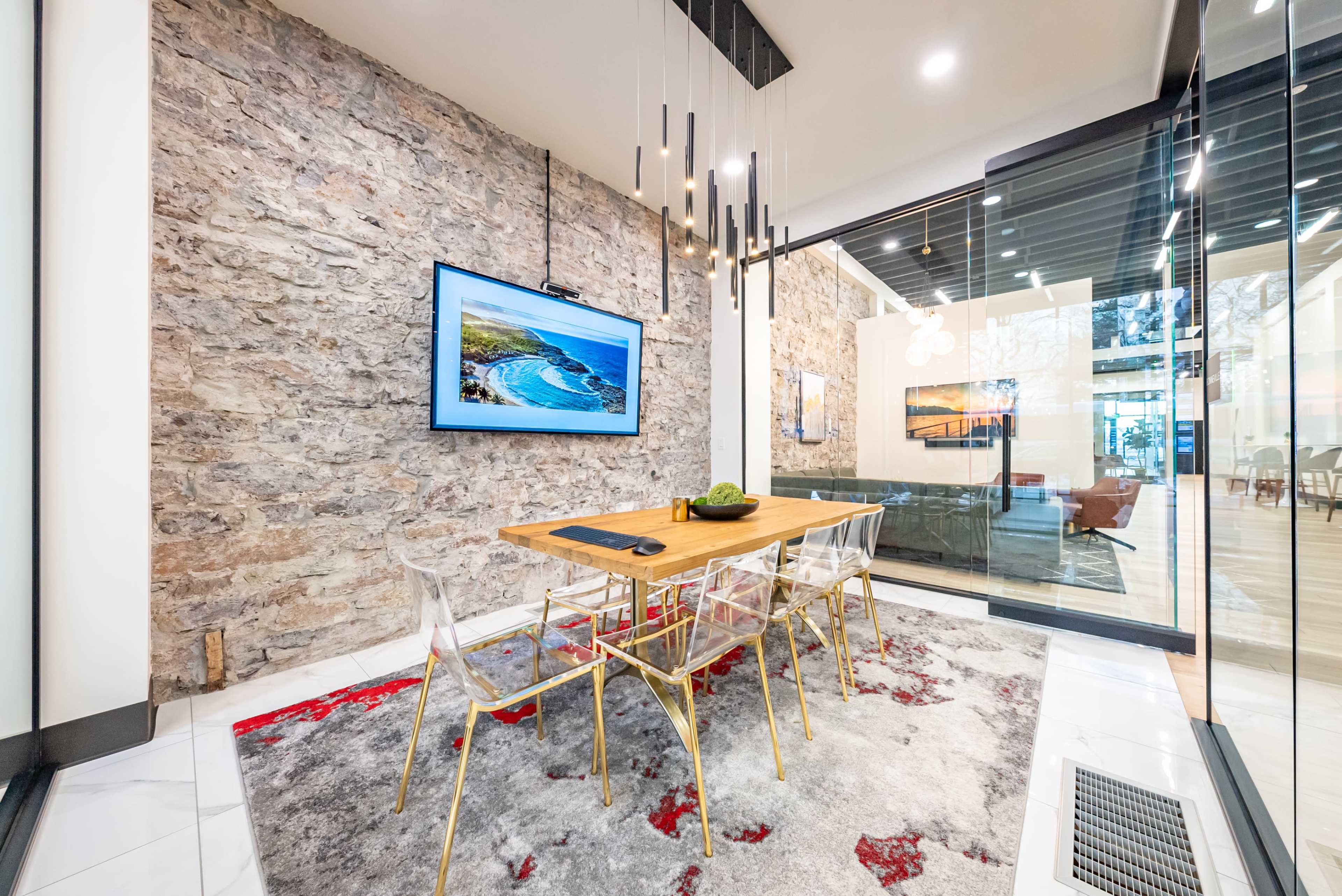A modern dining area with a wooden table surrounded by transparent chairs, a large TV mounted on a stone wall, and large glass panels that open to a spacious interior.