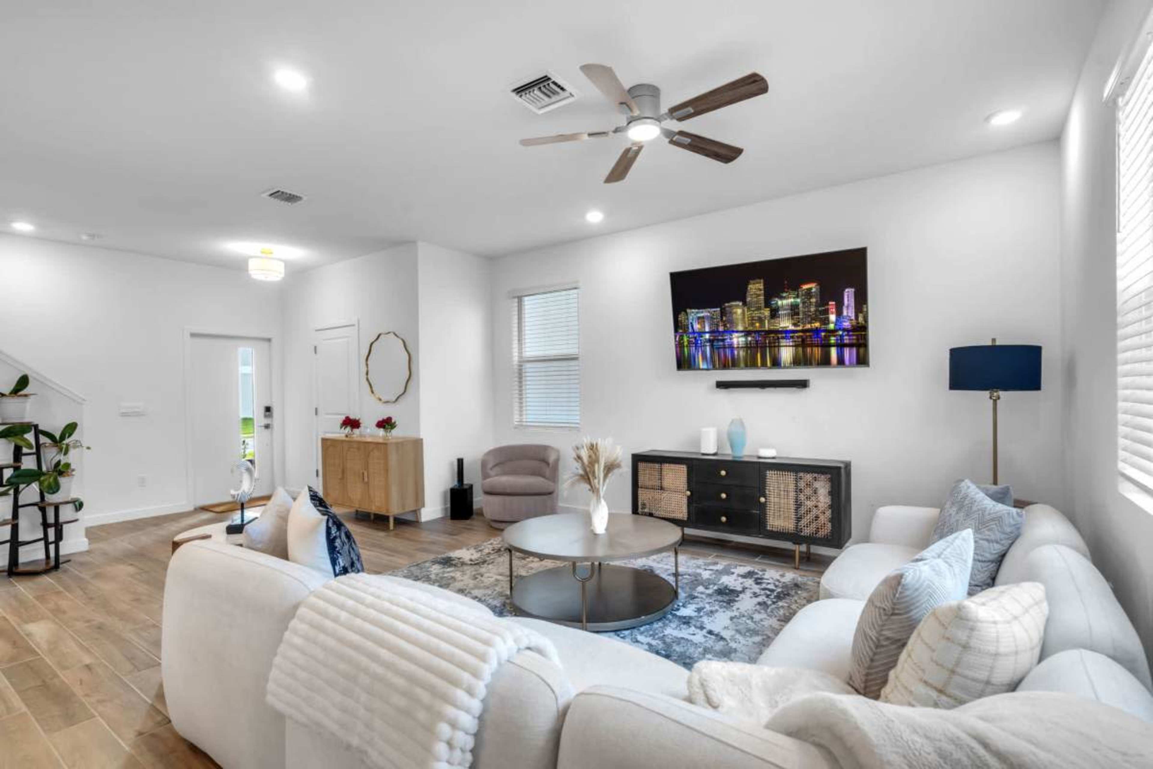 The image shows a modern living room featuring a white sectional couch, a round coffee table, a mounted television displaying a city skyline, and a wooden cabinet against the wall.
