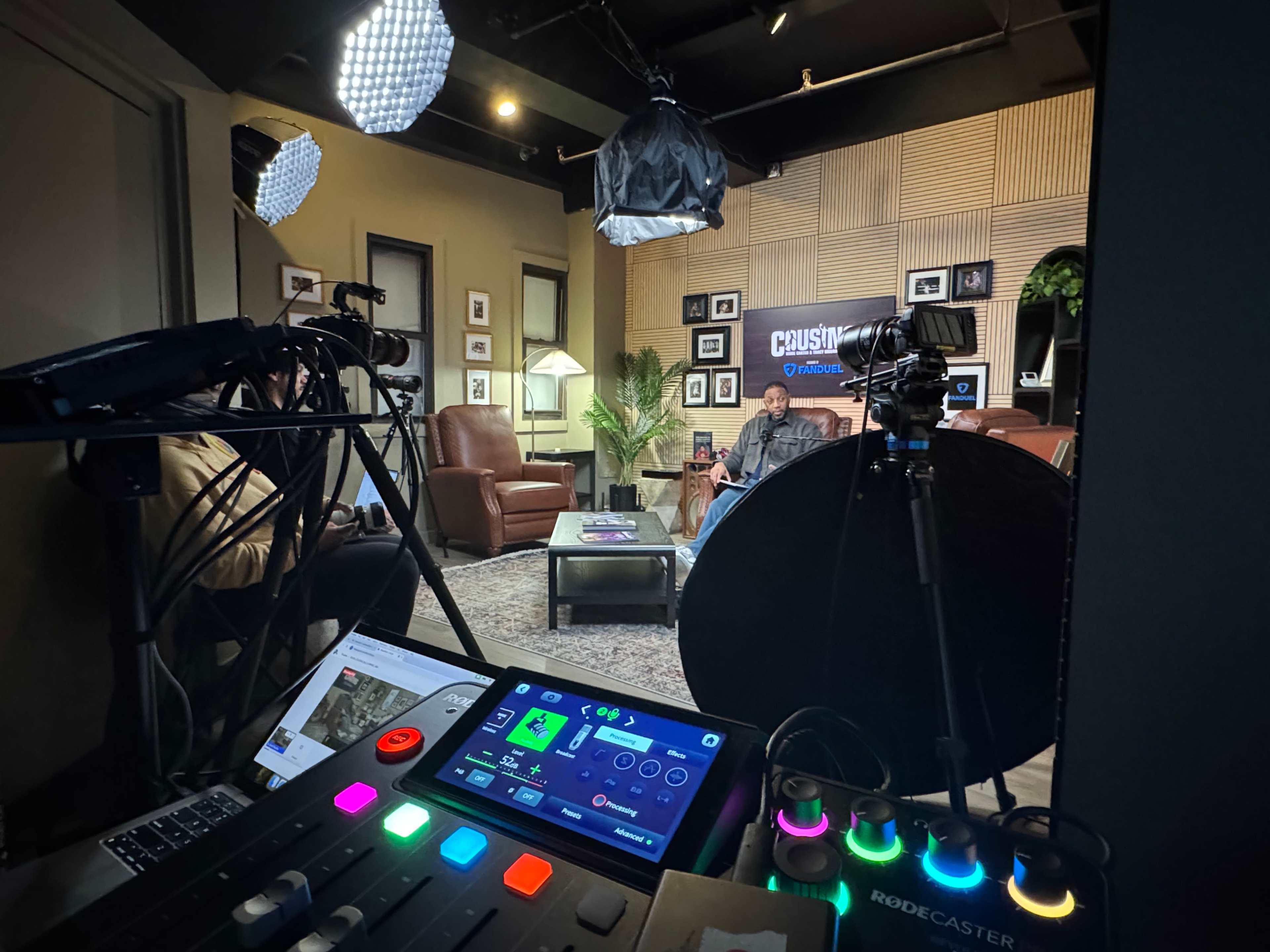The image shows a recording studio setup with a person seated in a lounge area, surrounded by cameras, lighting equipment, and sound mixing gear.