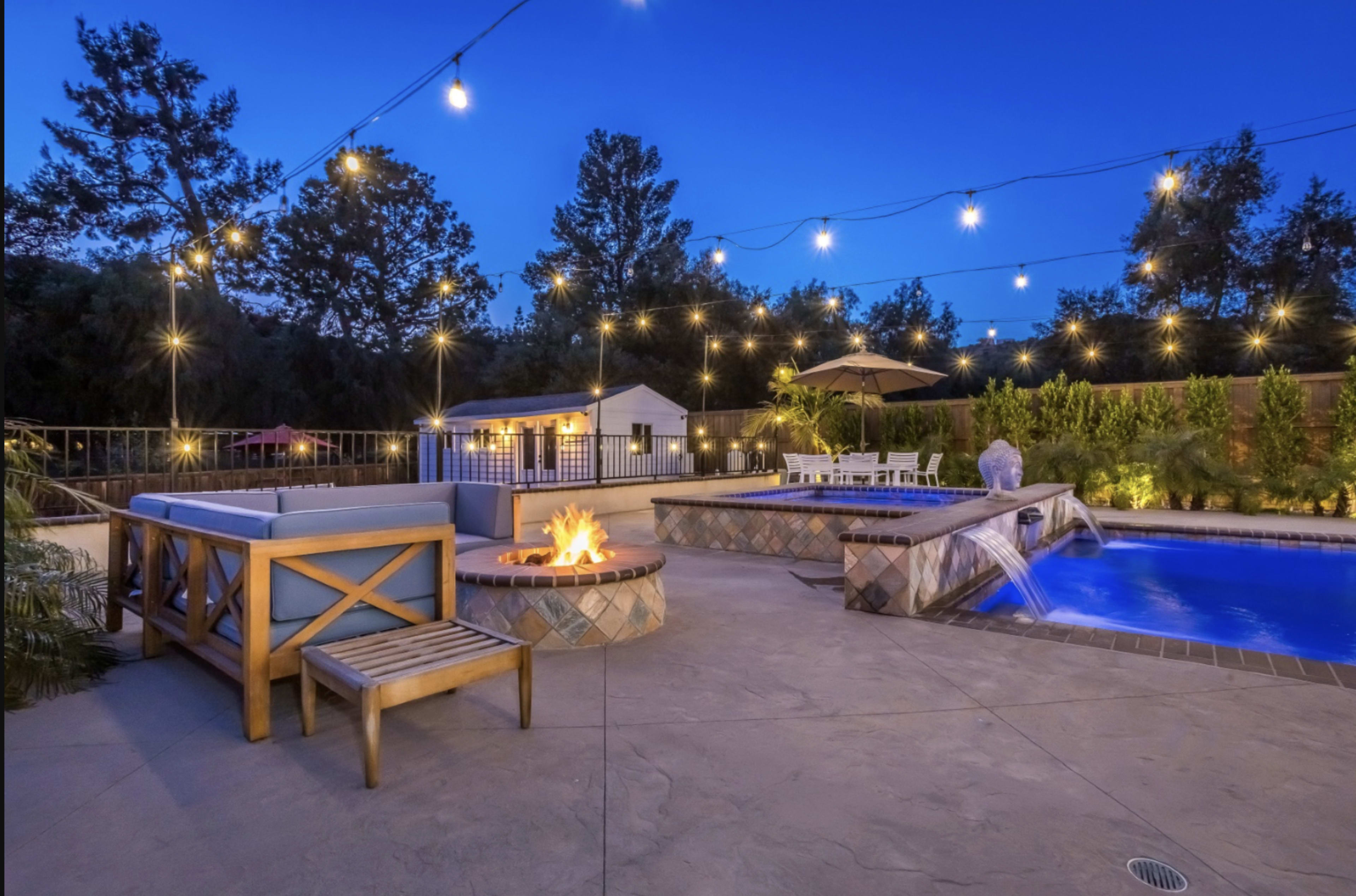 A well-lit outdoor area features a swimming pool, a fire pit with seating, and lounge spaces, surrounded by trees and a clear evening sky.