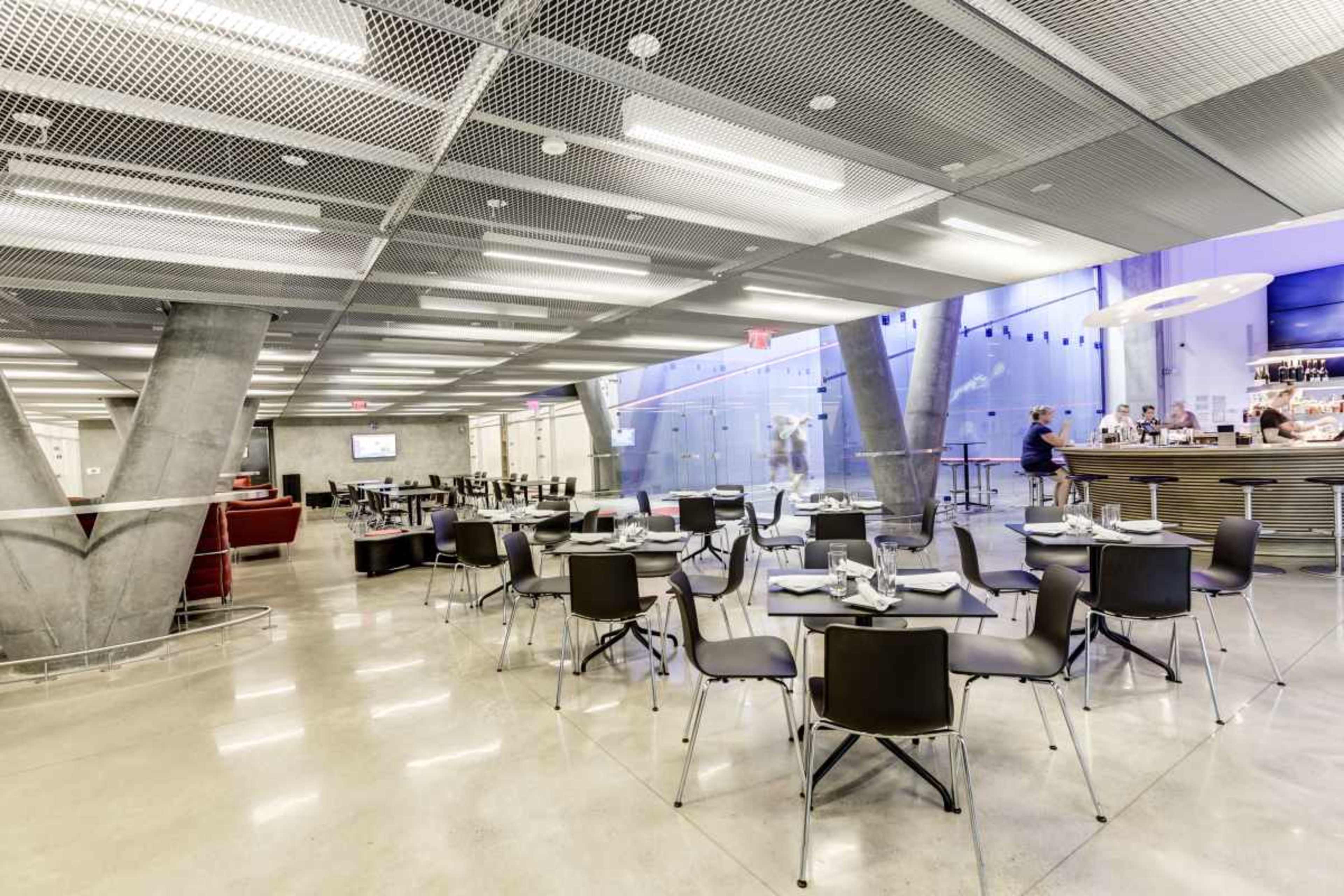 The image shows a modern restaurant interior with metal and concrete elements, featuring multiple tables and chairs arranged for dining, along with a bar area in the background.