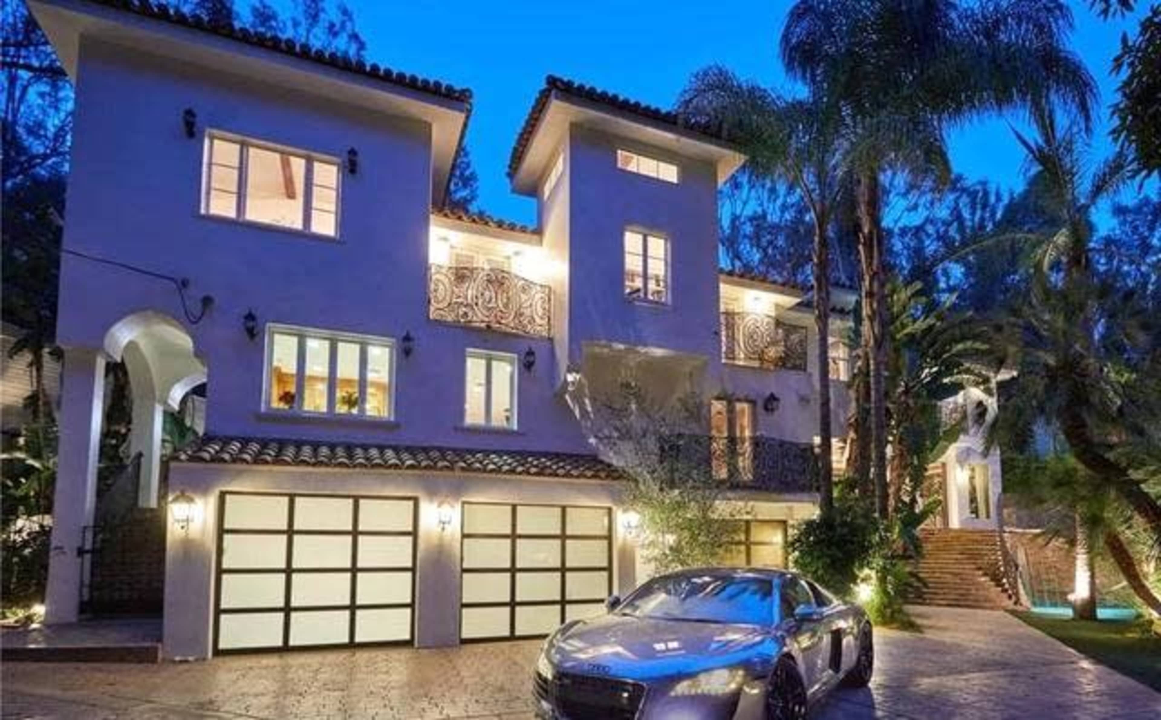 A modern, two-story house with a tile roof and multiple balconies is illuminated at dusk, accompanied by a luxury car parked in the driveway.