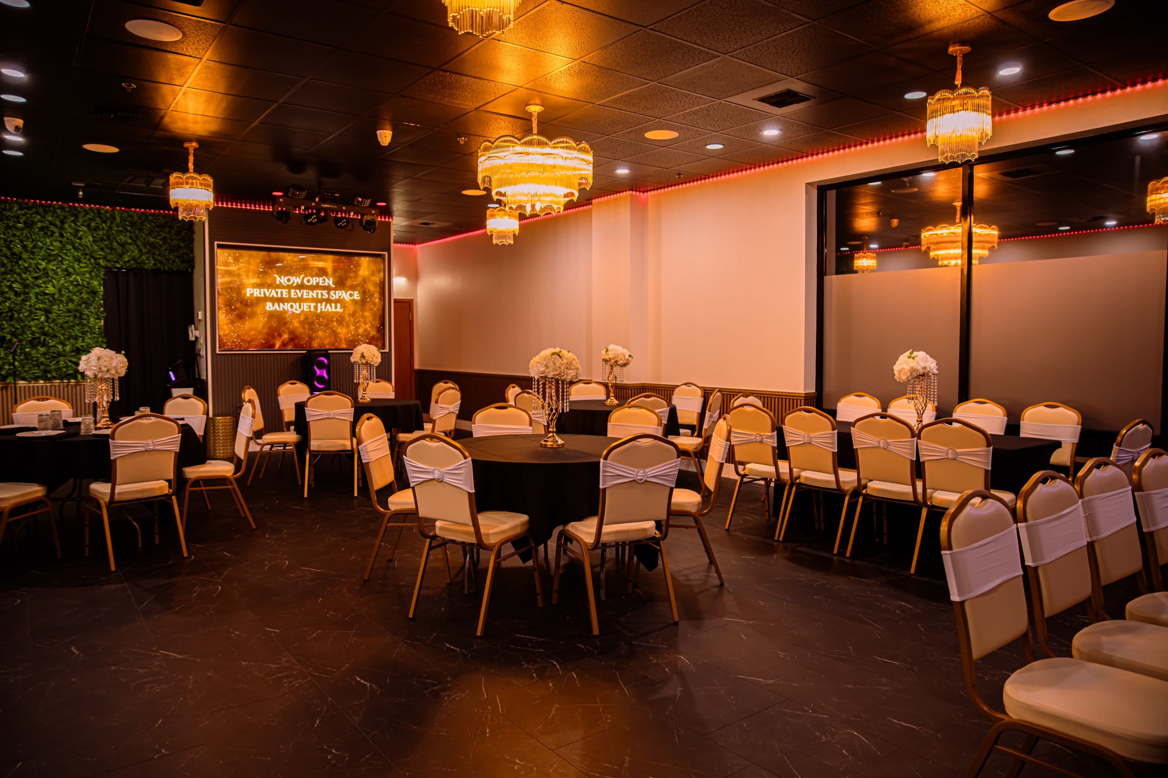 The image shows a banquet hall with round tables covered in black tablecloths, surrounded by gold and white chairs, and featuring chandeliers and a lit-up sign at the front.