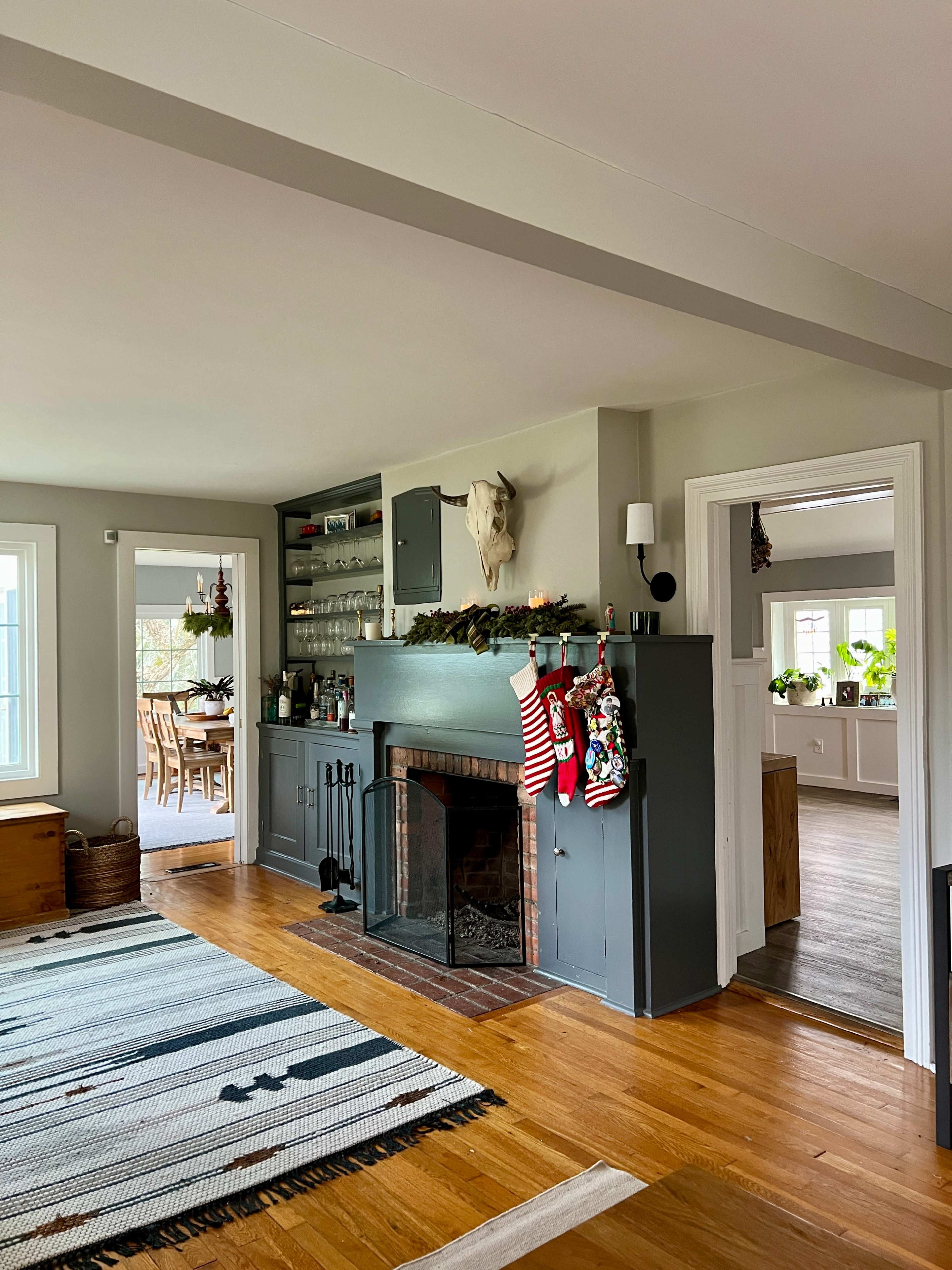 The image shows a living room featuring a fireplace with stockings hung on a gray mantel, surrounded by wooden flooring and an adjoining dining area visible in the background.