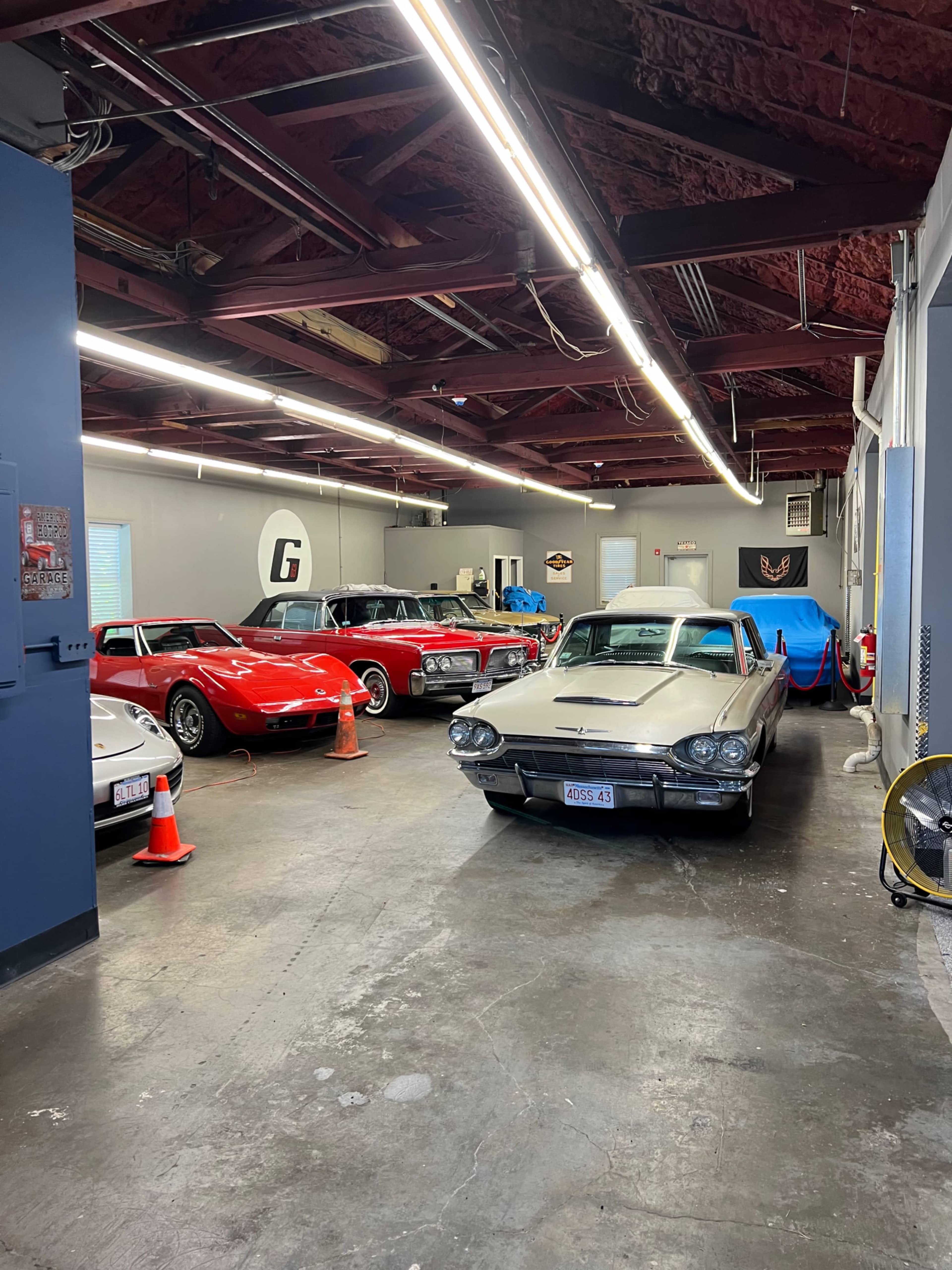 A spacious garage with several classic cars, including a silver Thunderbird and a red Corvette, parked under bright overhead lights.