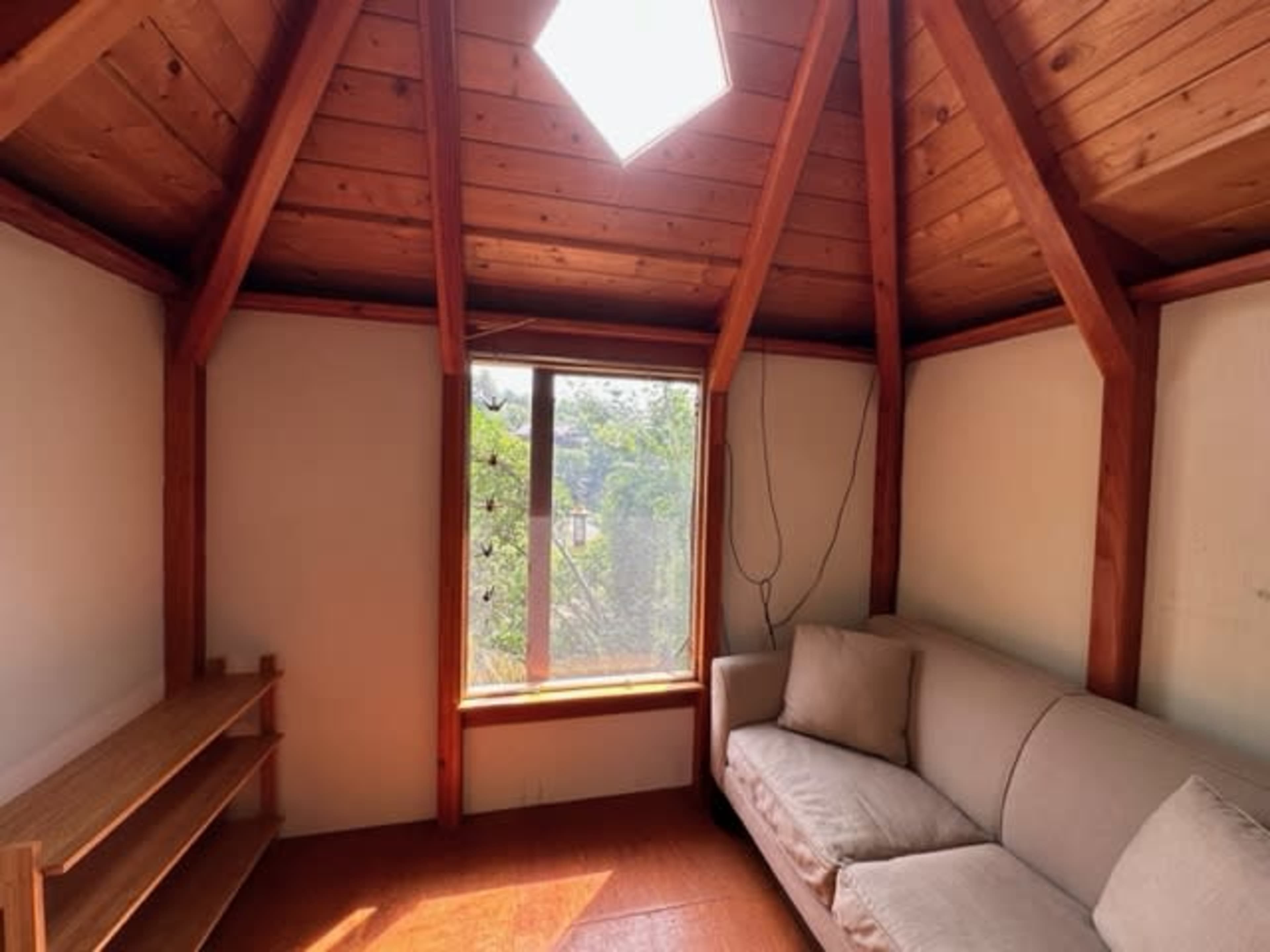 The image shows a small, wooden cabin interior with a sofa and a large window allowing natural light to enter.