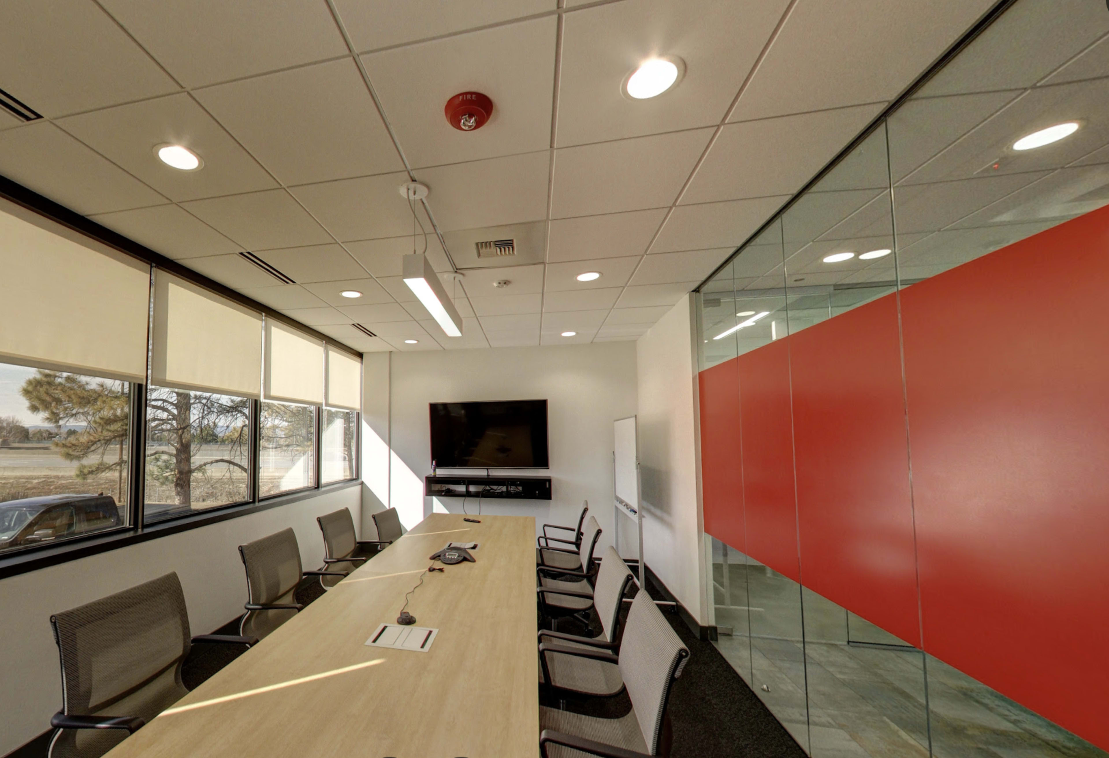 The image depicts a modern conference room with a long wooden table, office chairs, a wall-mounted television, and a glass partition featuring a red accent wall.