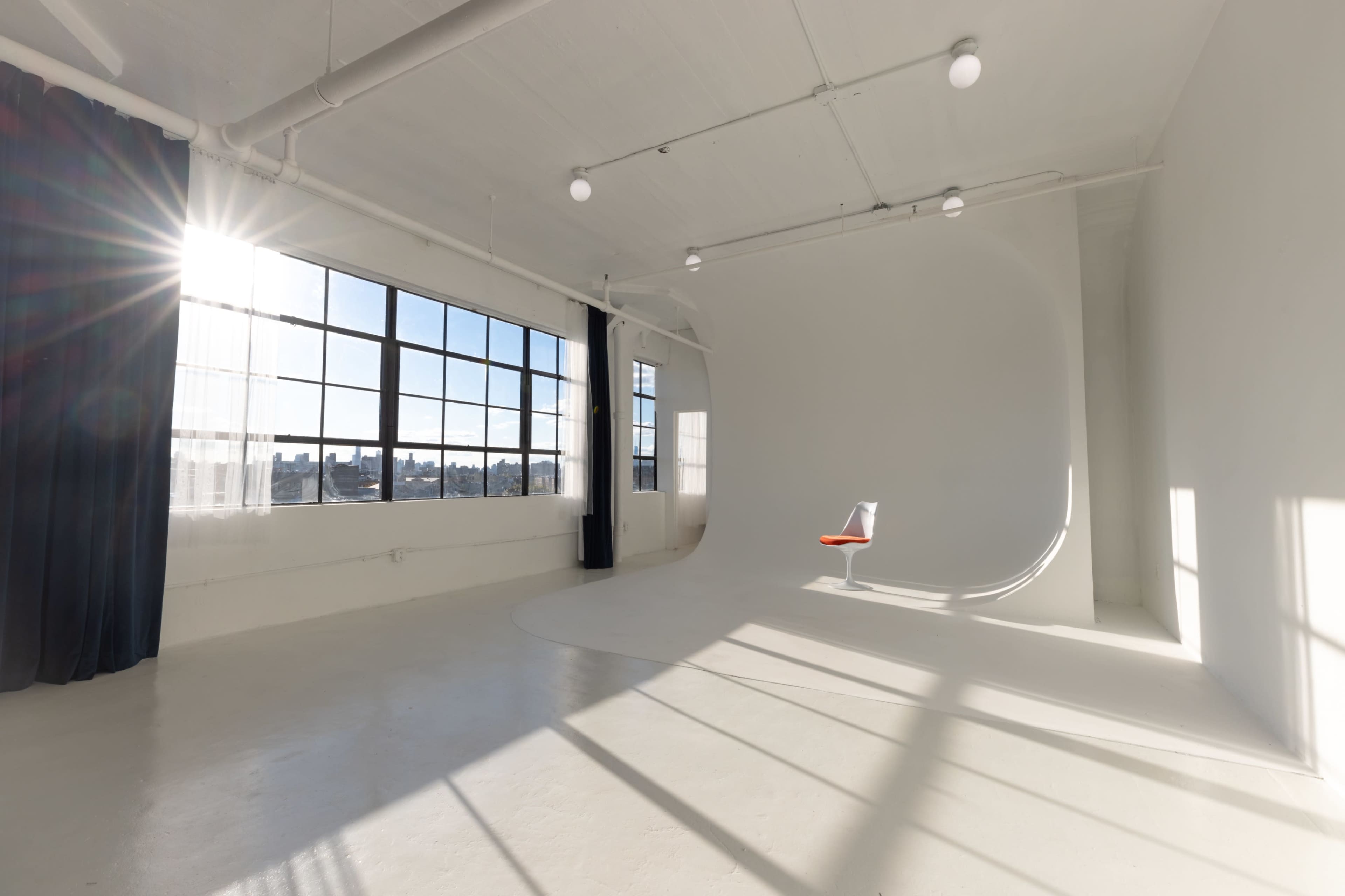 A bright, spacious studio features a curved white backdrop, a single chair with an orange seat, and large windows letting in sunlight.