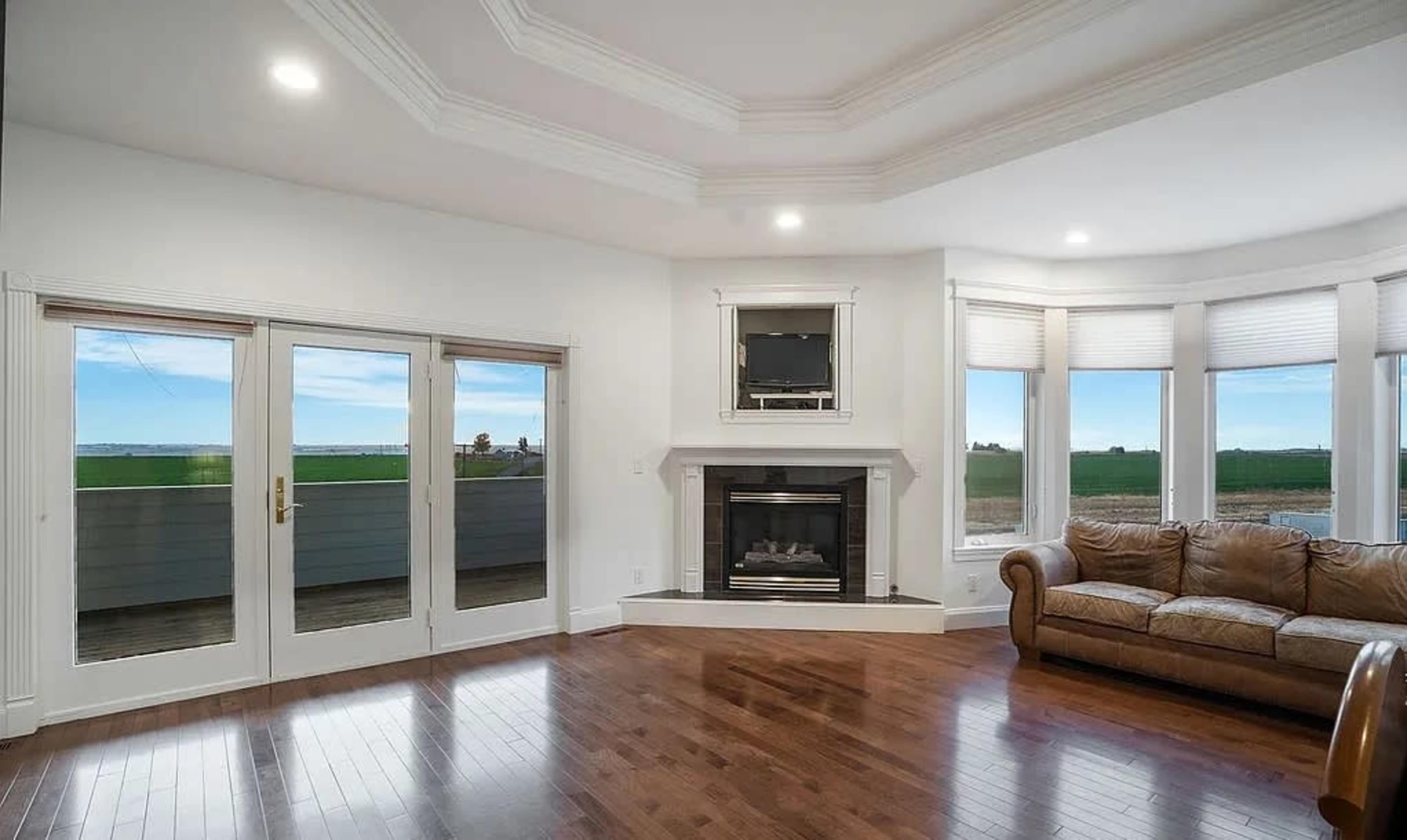 The image shows a living room with hardwood floors, a fireplace, large windows, and a leather sofa, offering a view of the outdoors.