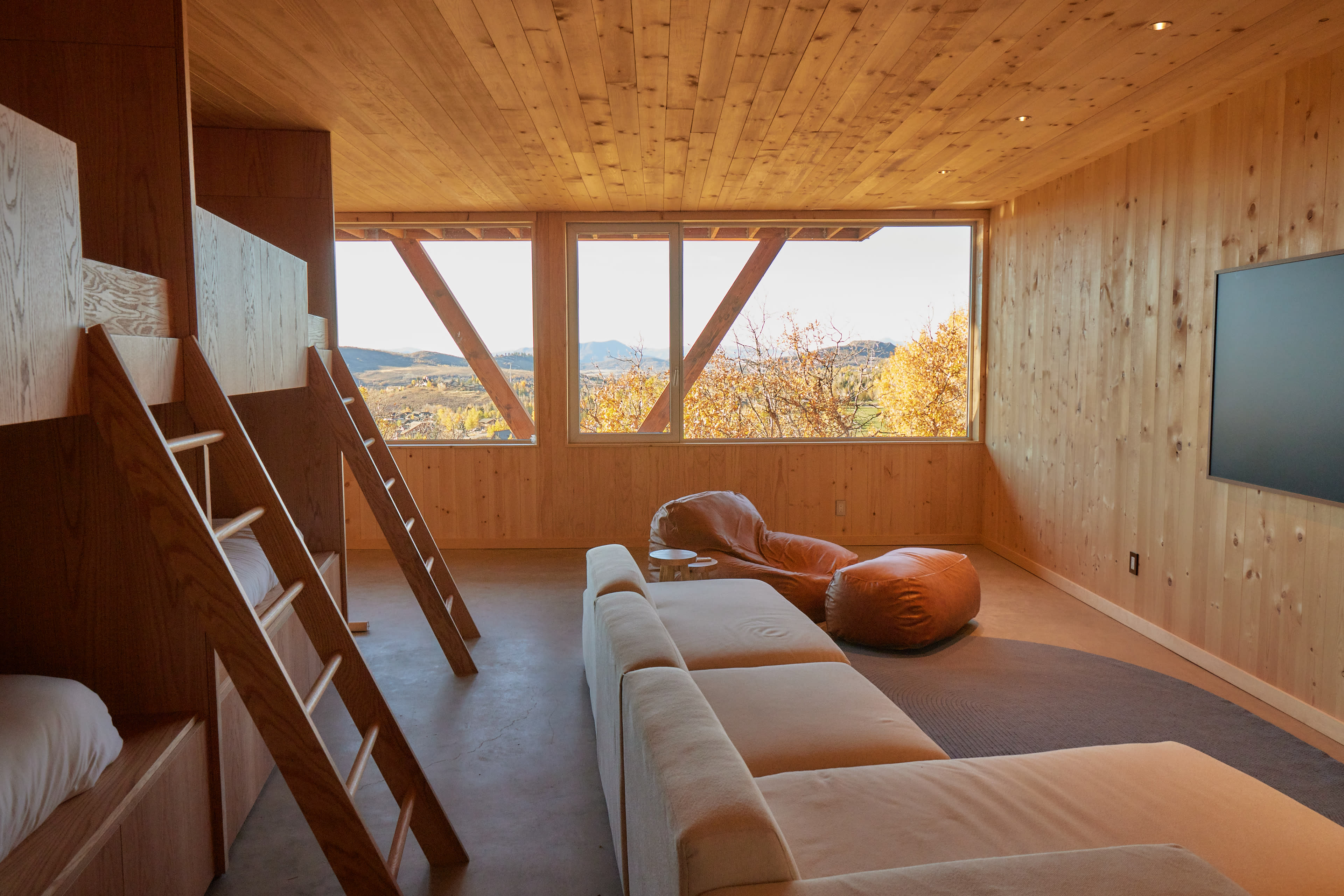 The image shows a modern wooden interior of a room featuring a sofa, a ladder leading to a lofted bed, and large windows with a view of a natural landscape.