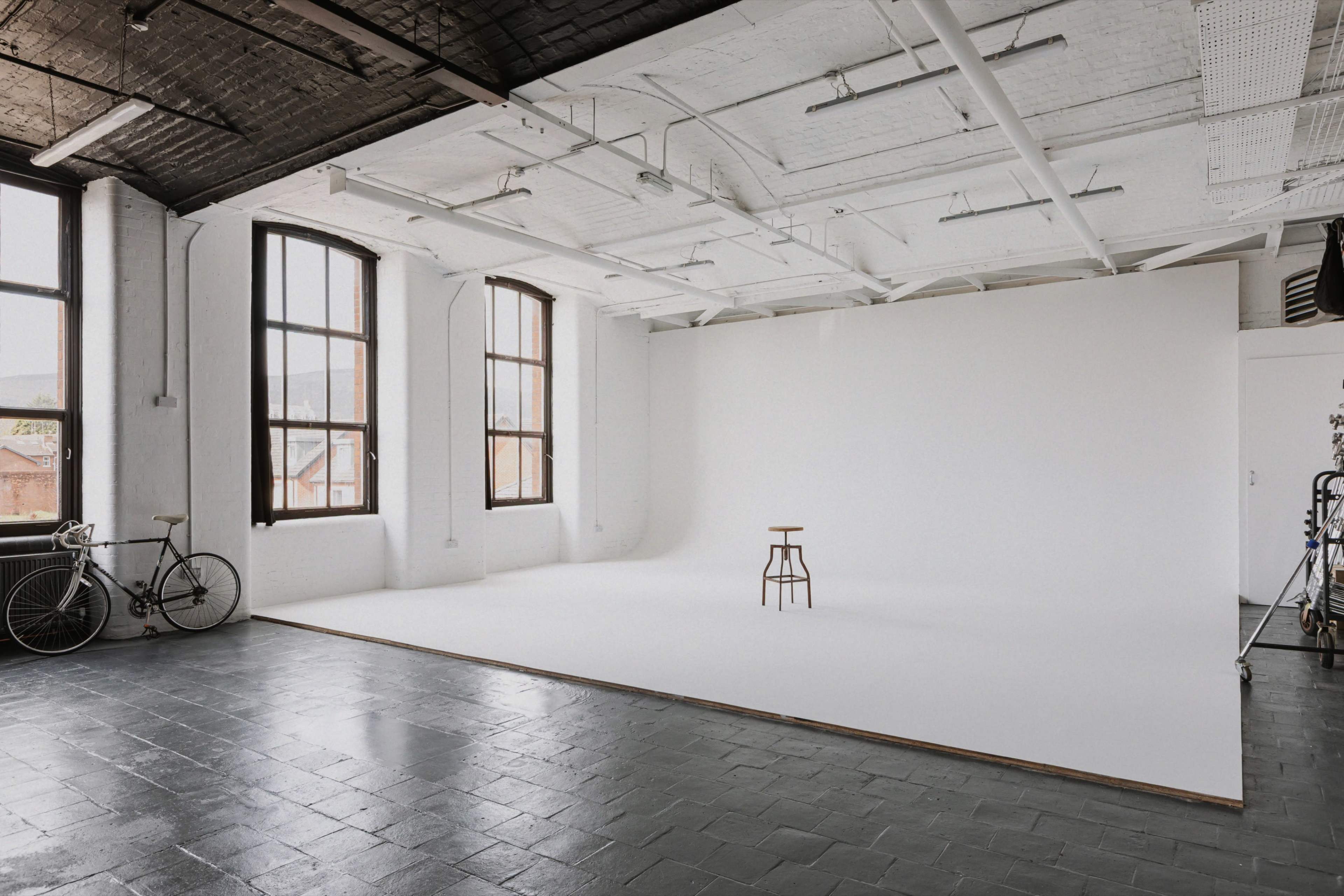 The image depicts a spacious studio with white walls, large windows, a wooden stool in the center, and a bicycle leaning against the wall.