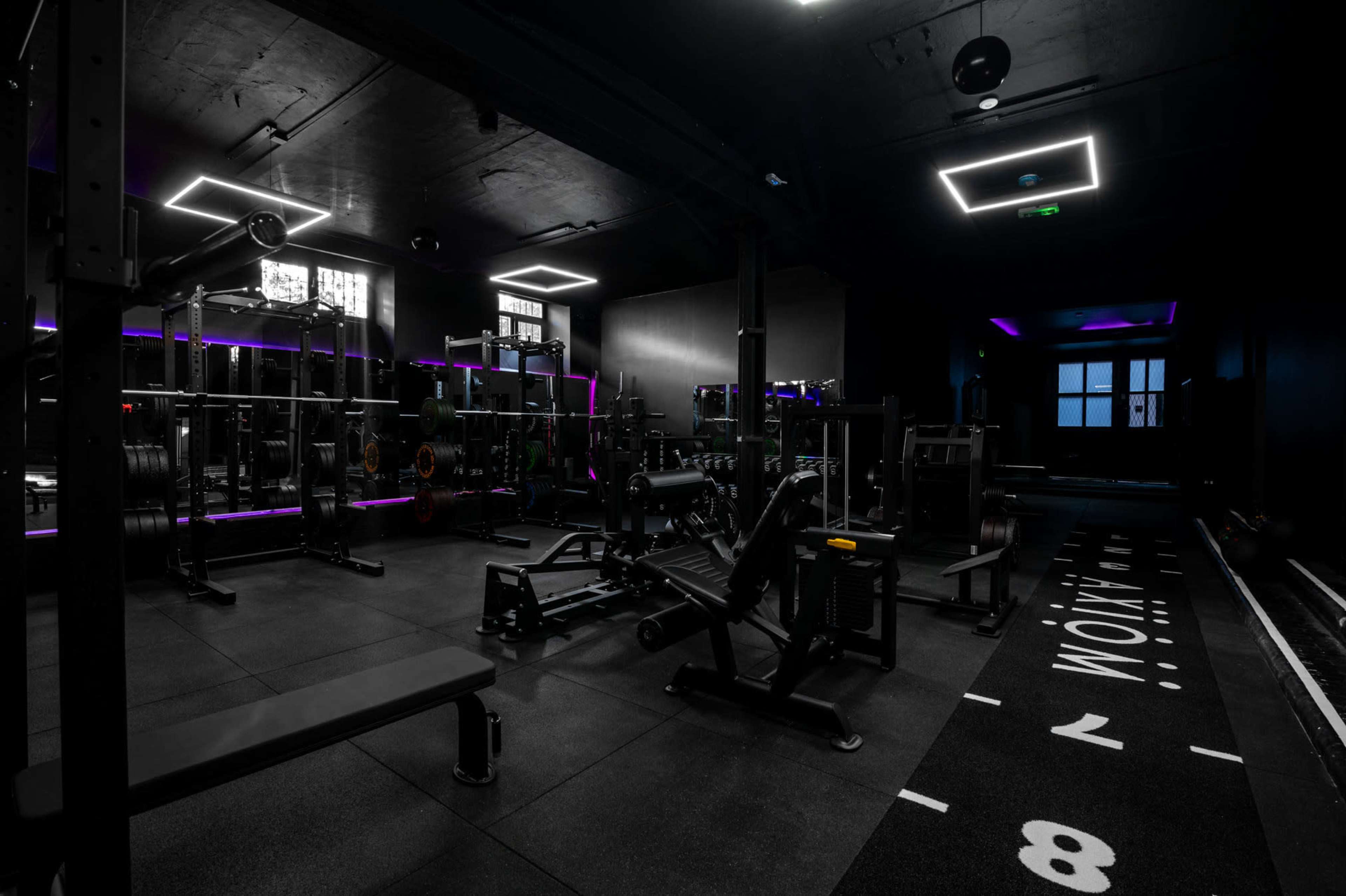 The image shows a dimly lit gym equipped with weightlifting benches, free weights, and exercise machines, with purple accent lighting and a black floor mat marked with numbers.