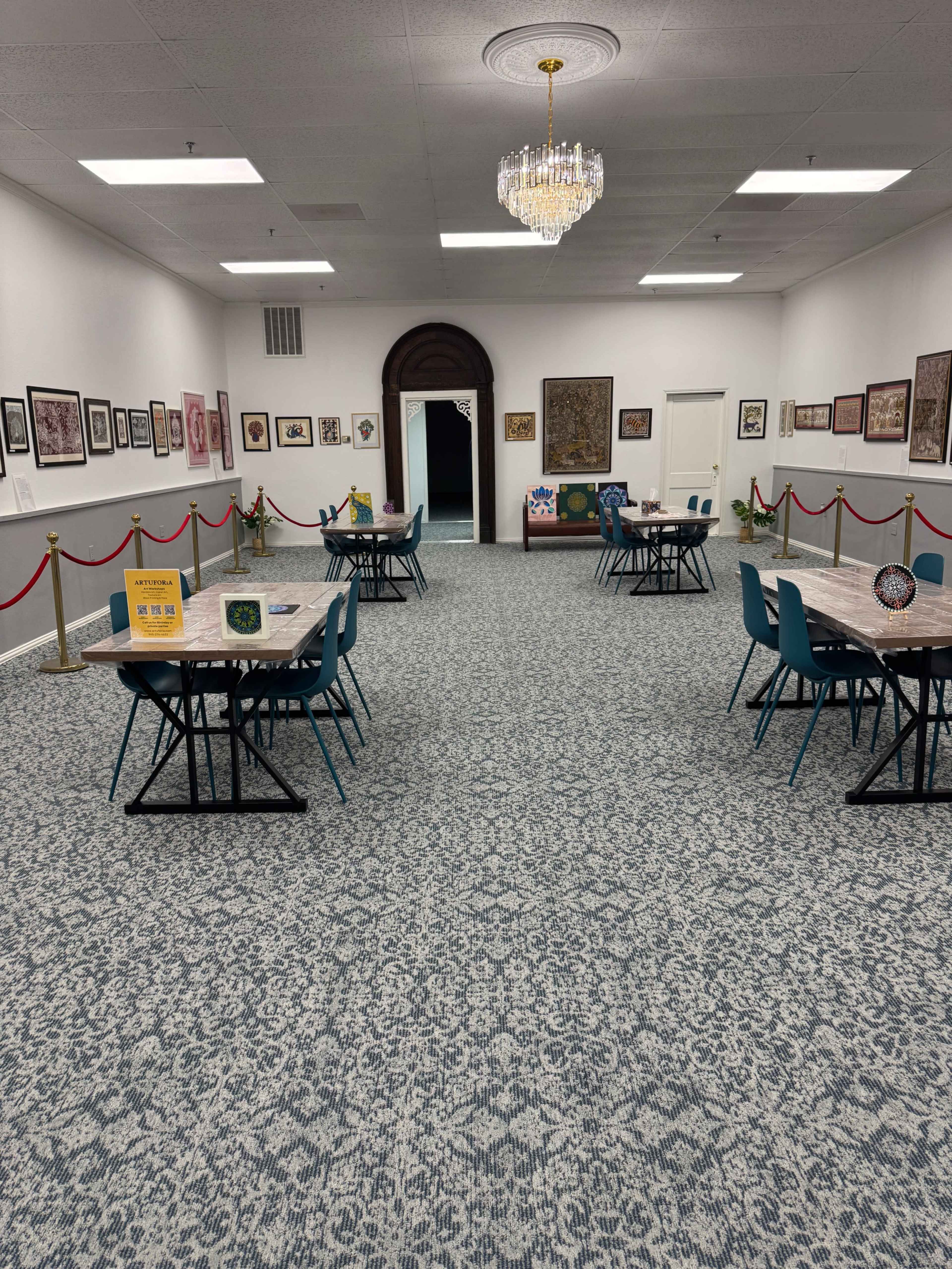 The image shows a spacious room with several tables and chairs arranged in a symmetrical layout, adorned with framed artwork on the walls and a chandelier hanging from the ceiling.