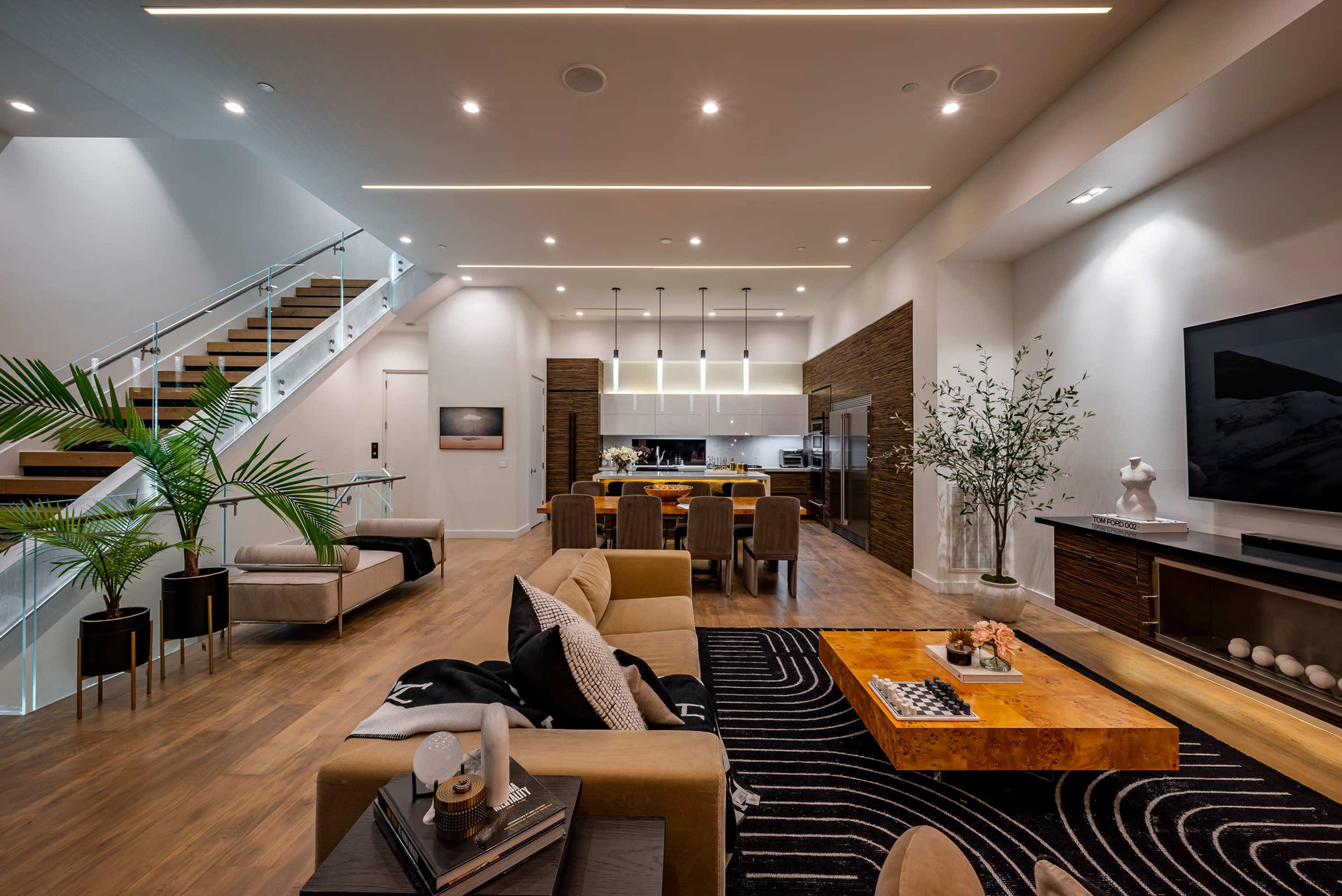 A modern living room and kitchen area featuring a staircase, large windows, a coffee table, and contemporary furnishings.