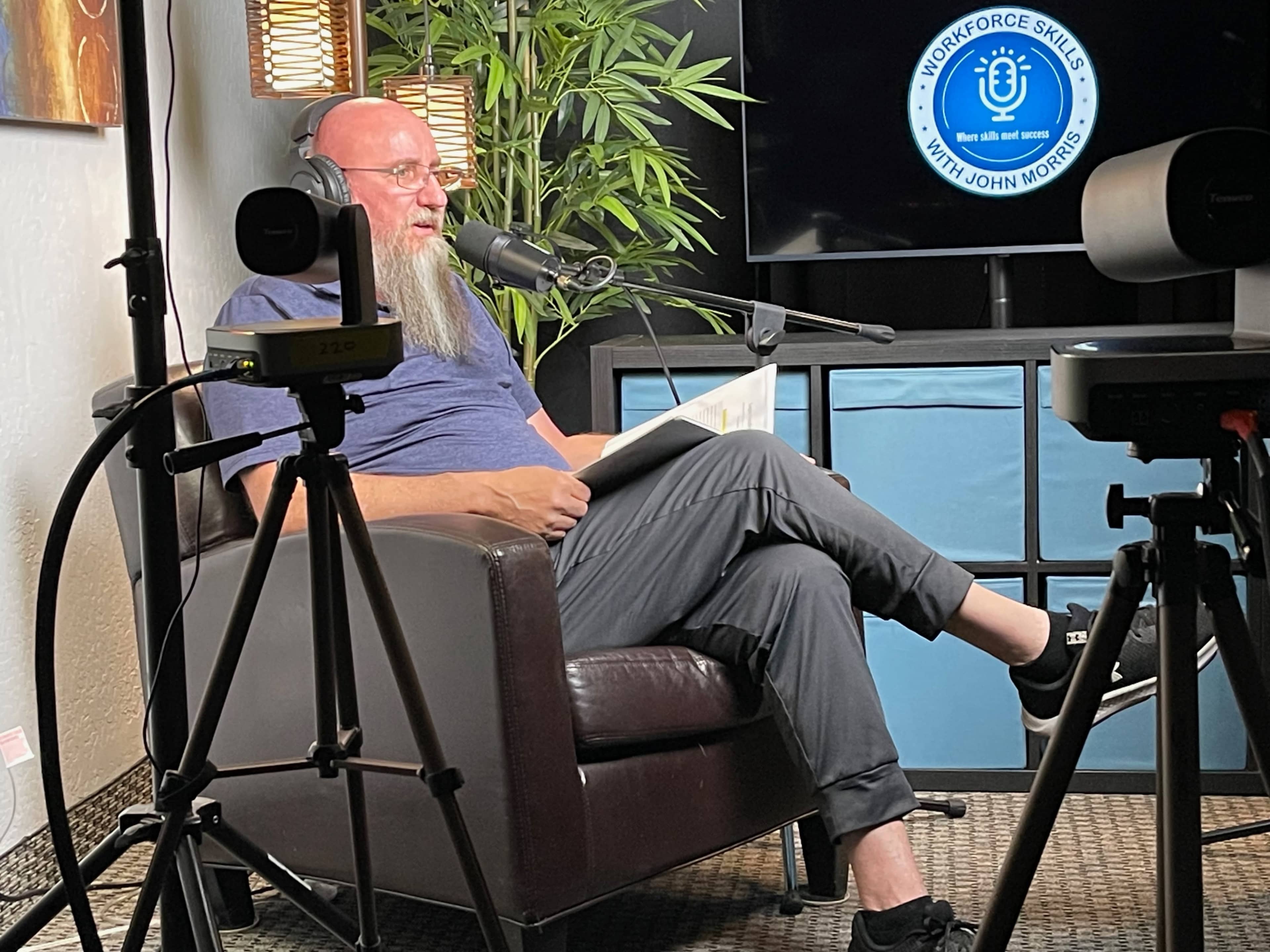 A man with a long beard sits in a chair reading from a document while speaking into a microphone, surrounded by cameras and a large screen in a well-lit room.