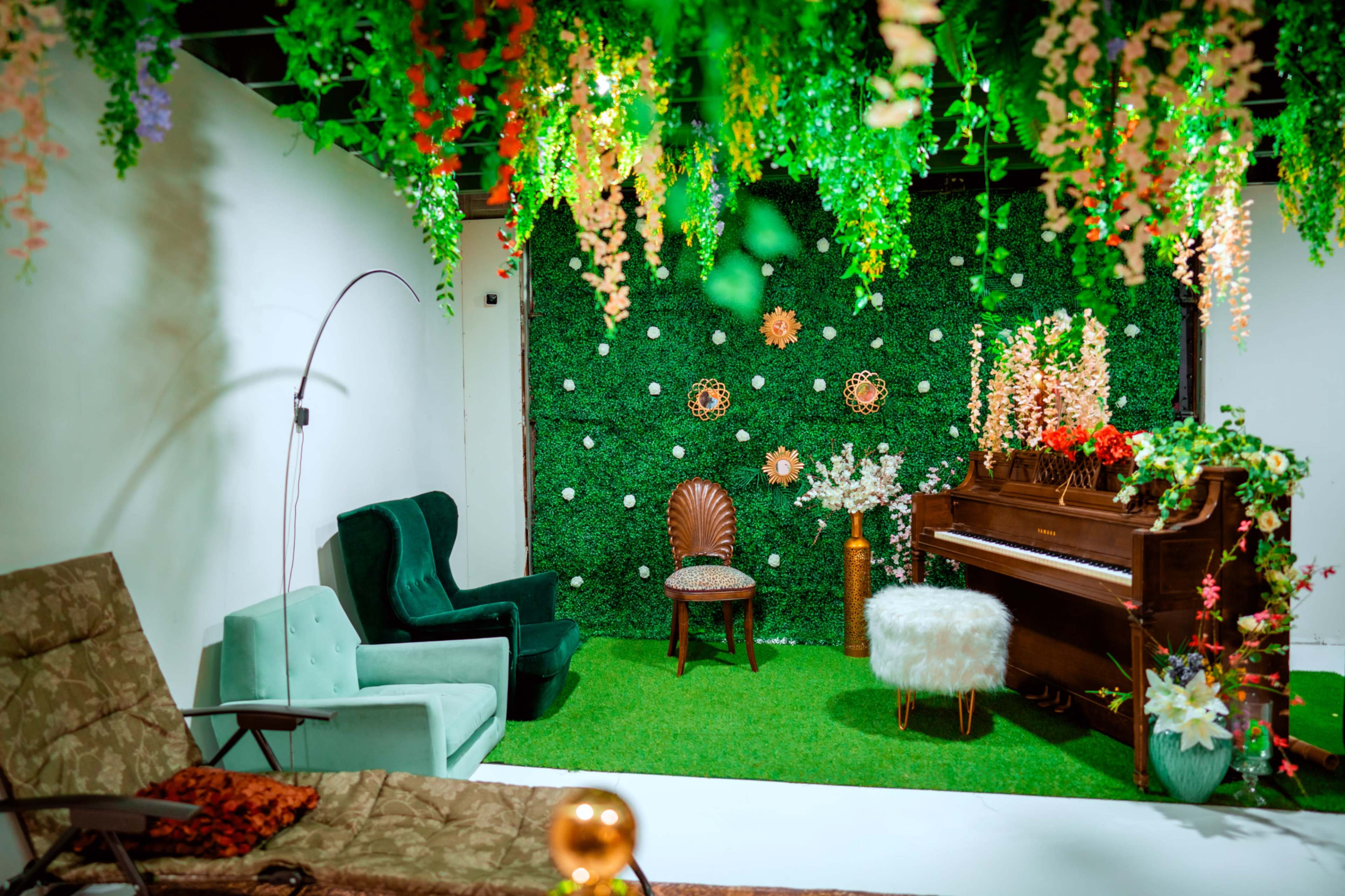 The scene features a decorated indoor space with artificial greenery, colorful flowers, a vintage piano, and a mix of chairs arranged on a grassy floor.