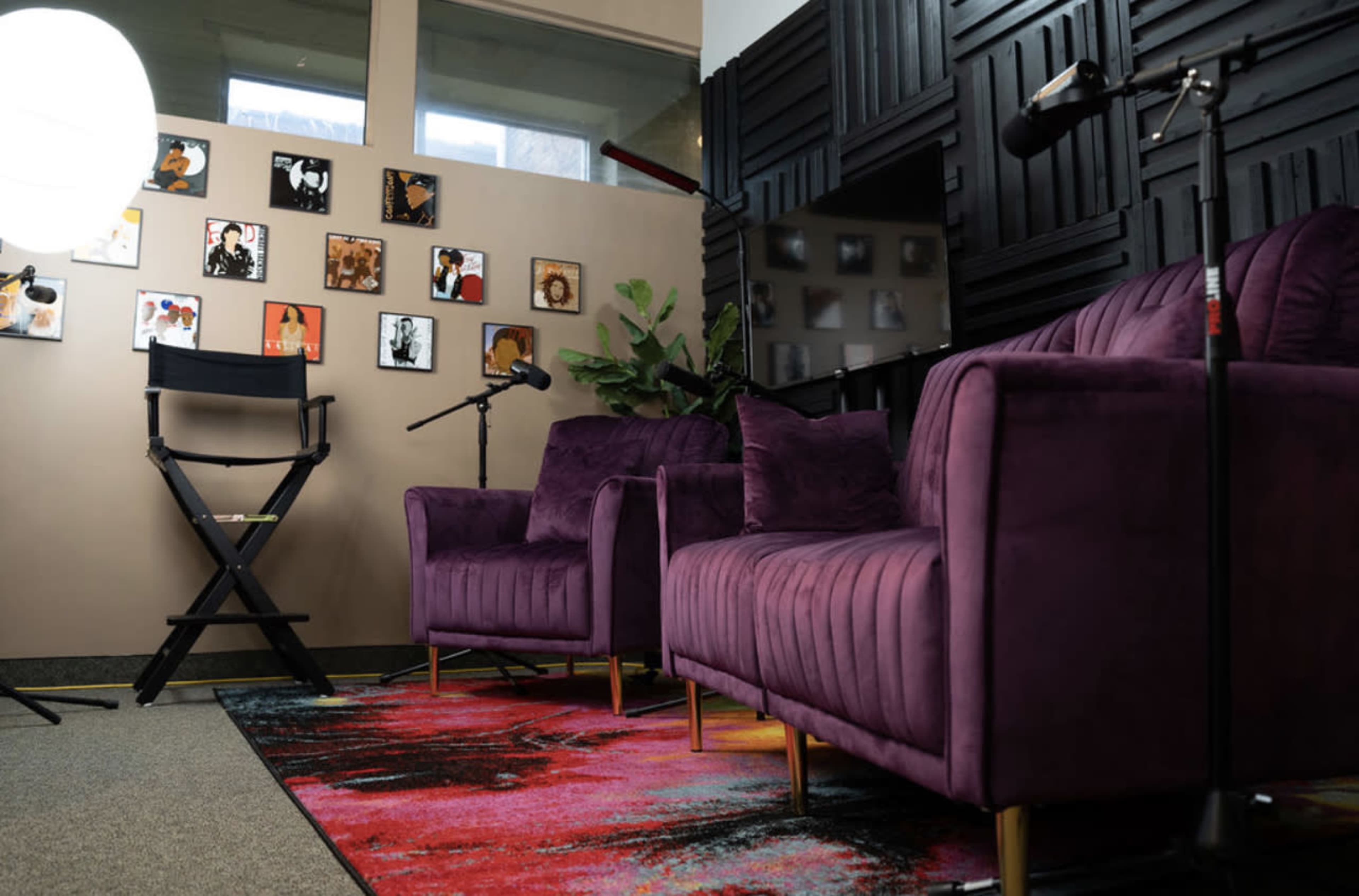 A small studio space featuring two purple chairs, a patterned rug, a director's chair, and several framed artworks on the wall.