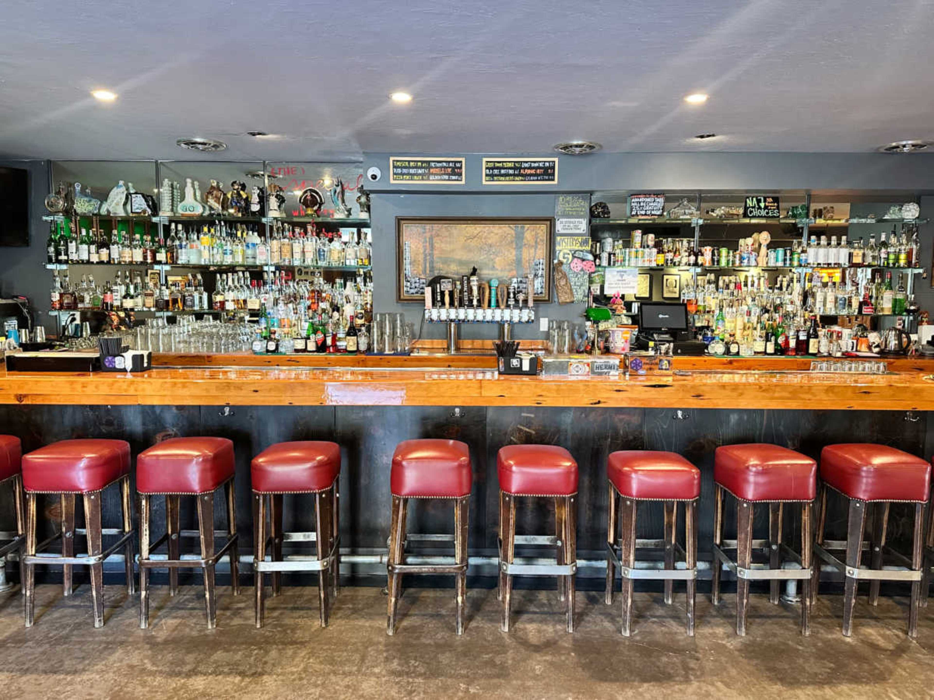 The image shows a well-stocked bar with a long wooden counter and red upholstered stools arranged in front.