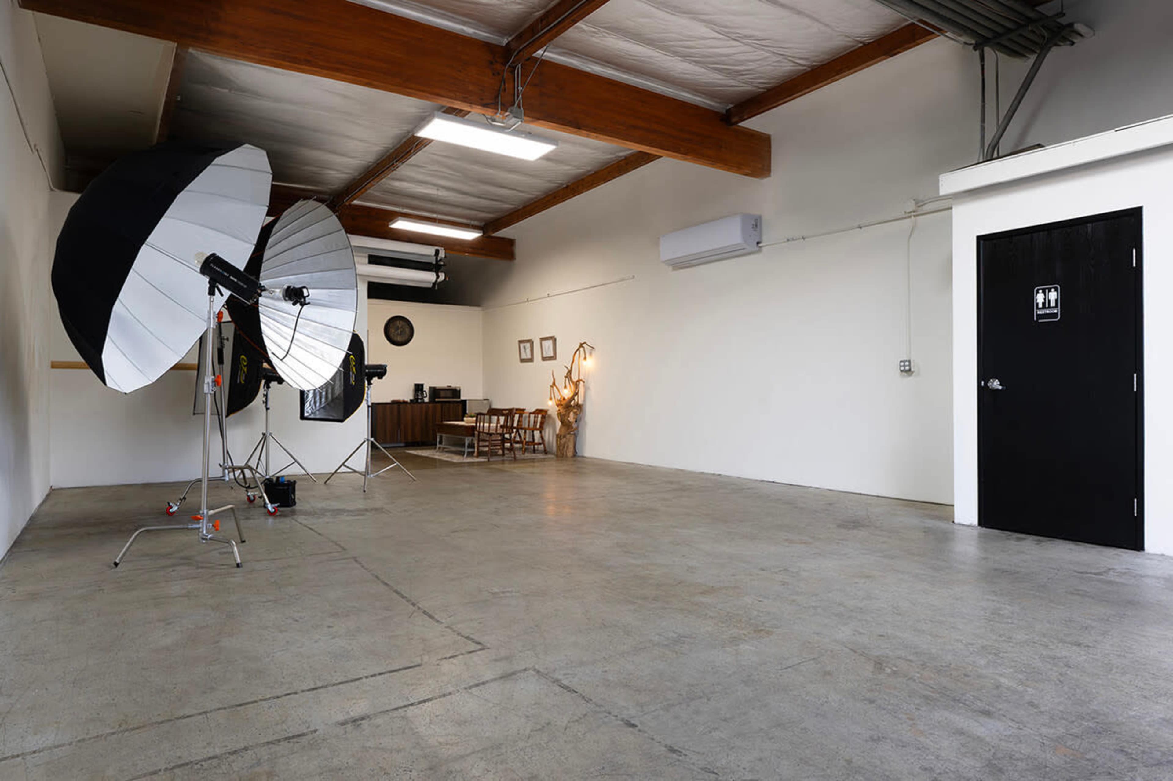 The image shows an empty studio space featuring large photography umbrellas, a desk with chairs, and a restroom door along a plain white wall.