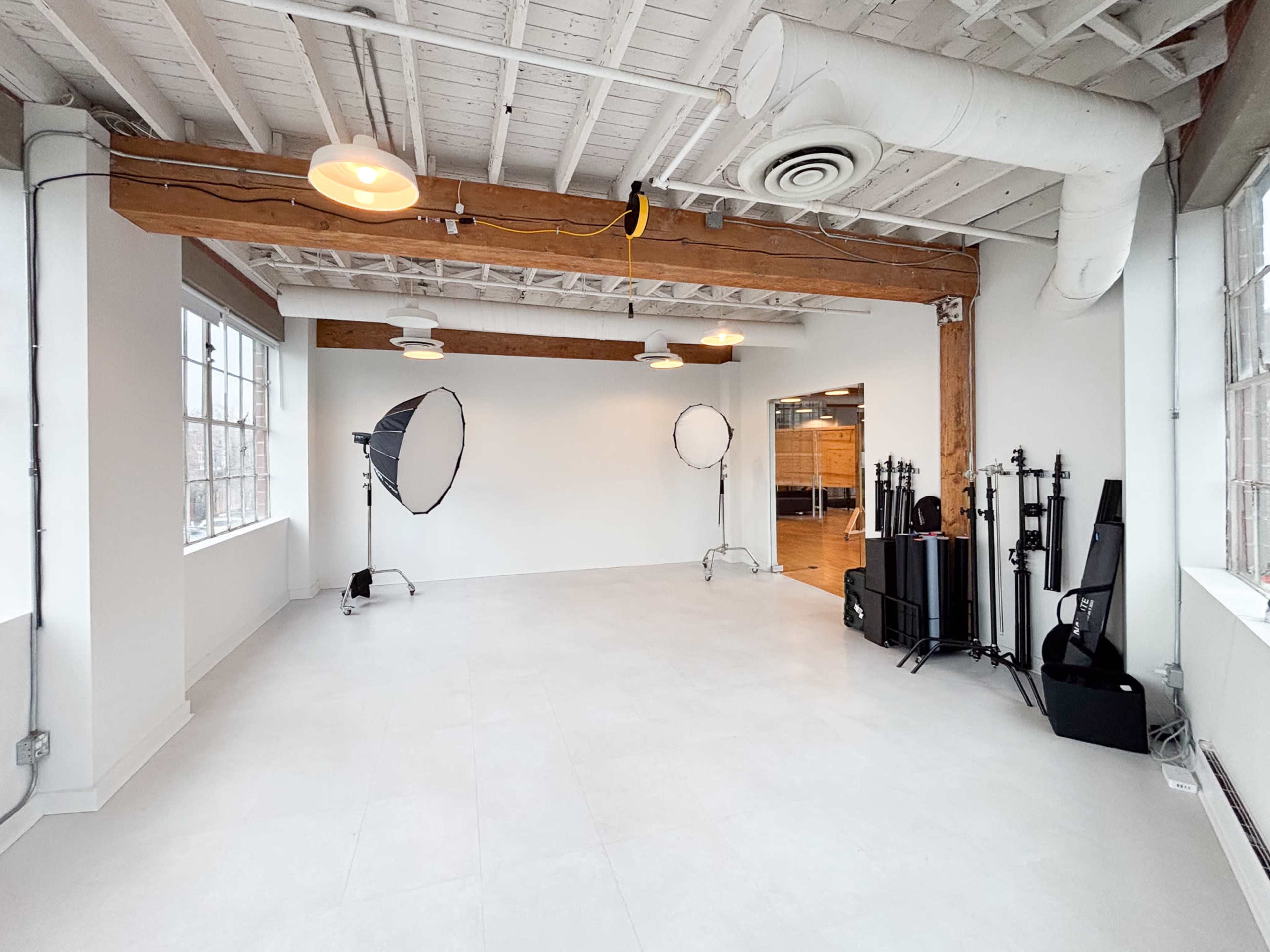 The image shows a spacious, empty studio with white walls, large windows, and studio lighting equipment set up on one side.