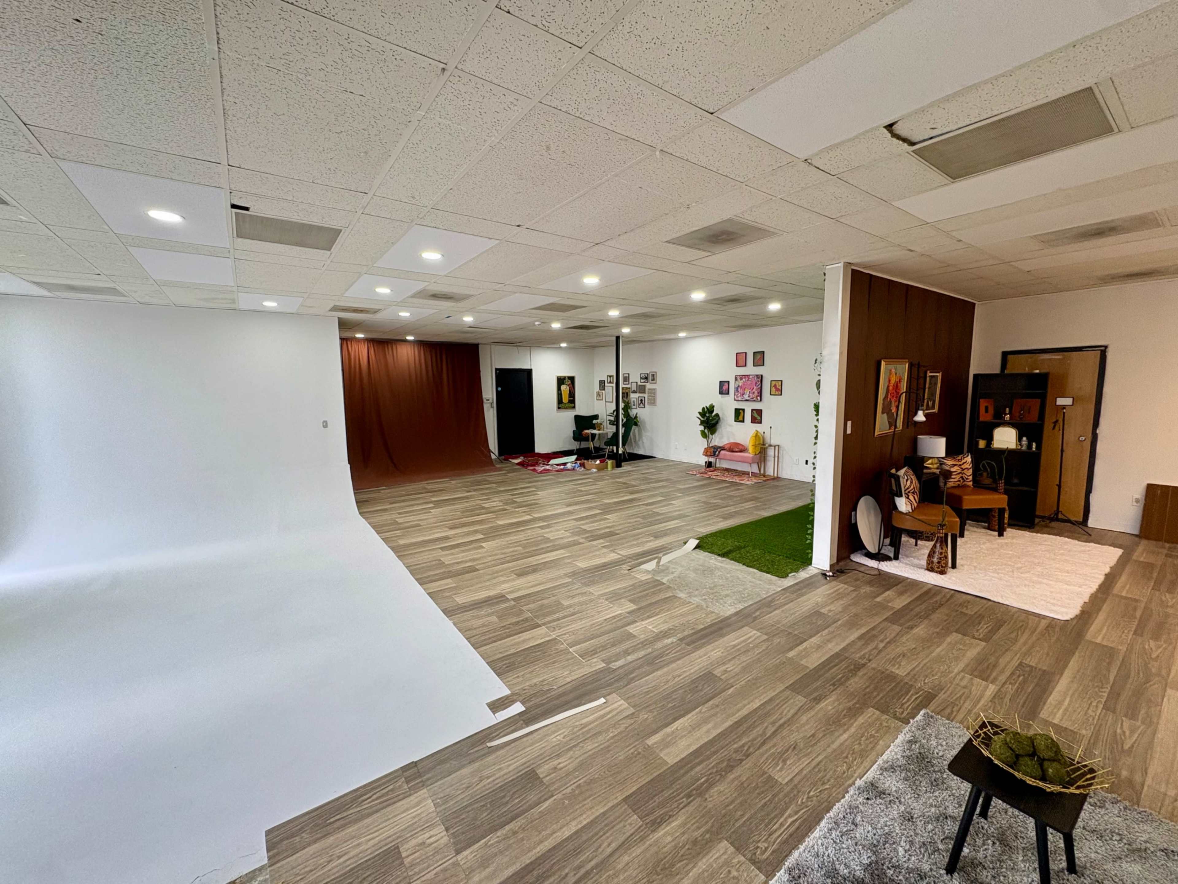 The image shows a spacious studio with a white backdrop on one side, wooden flooring, and a cozy sitting area featuring plants and artwork on the walls.