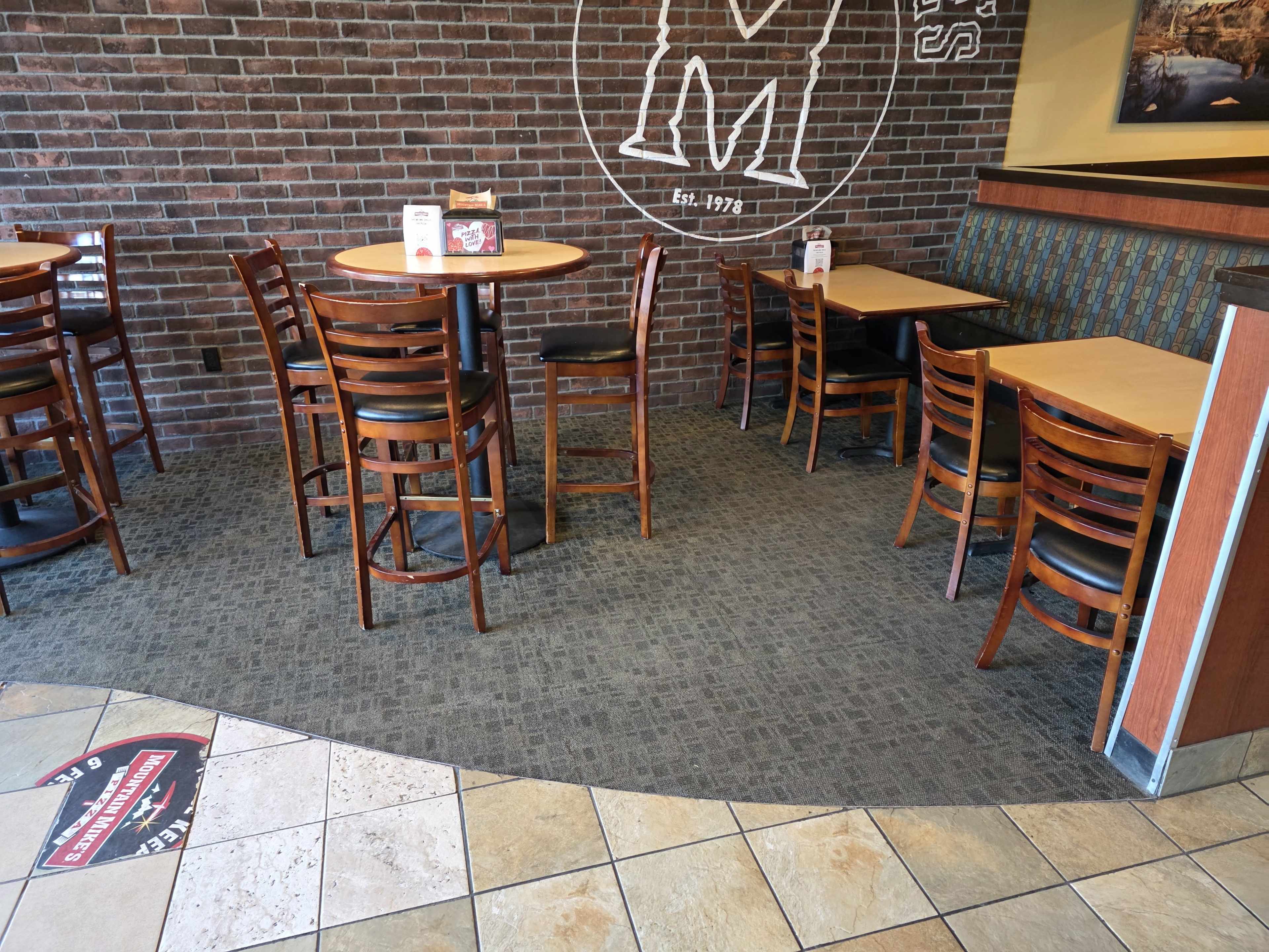 The image shows a corner of a restaurant with wooden tables and chairs arranged on a tiled floor near a brick wall.