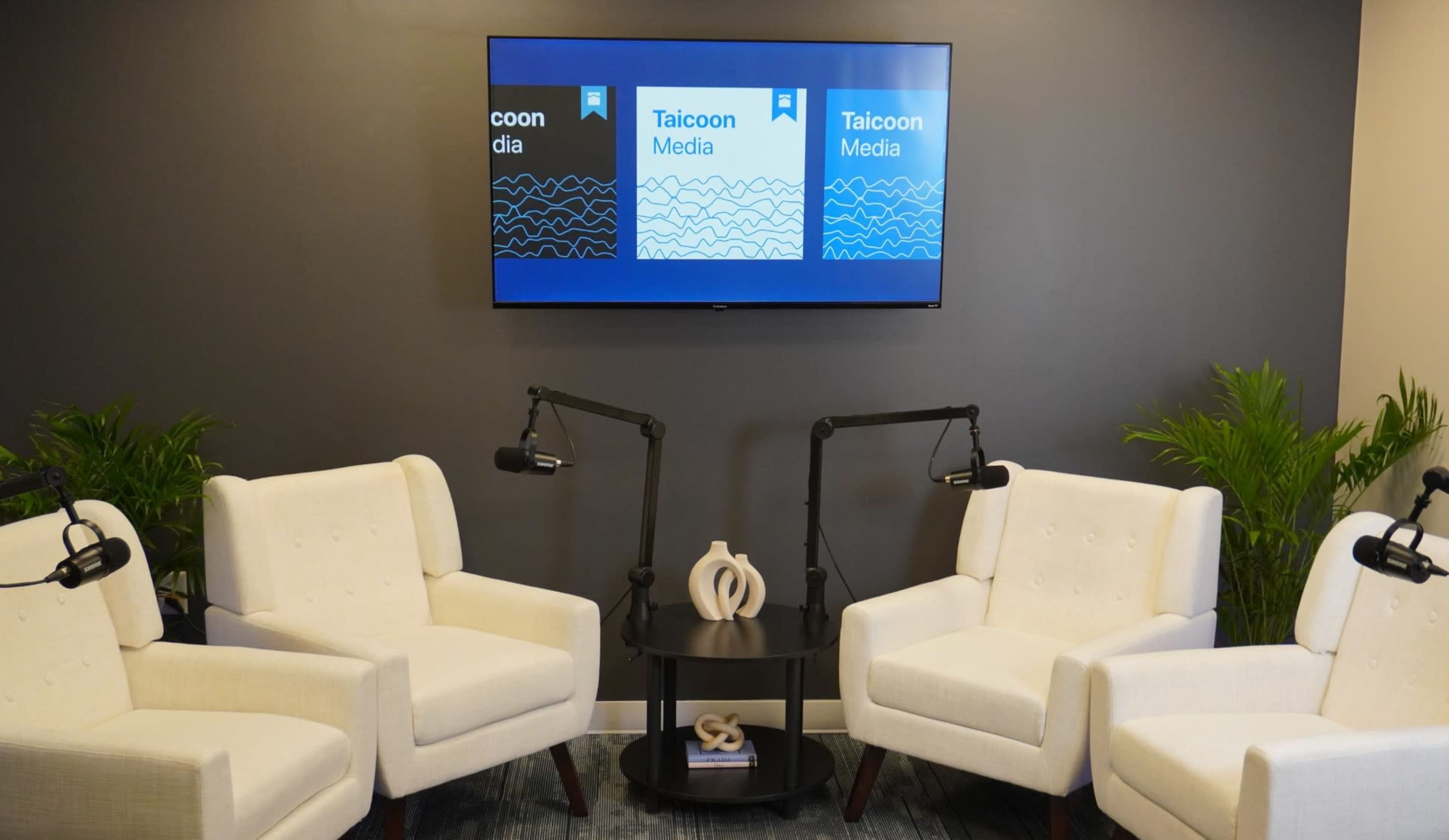The image shows a modern interview setup with two white armchairs, a small black table, and a television displaying the logo of Taicoon Media on a gray wall.