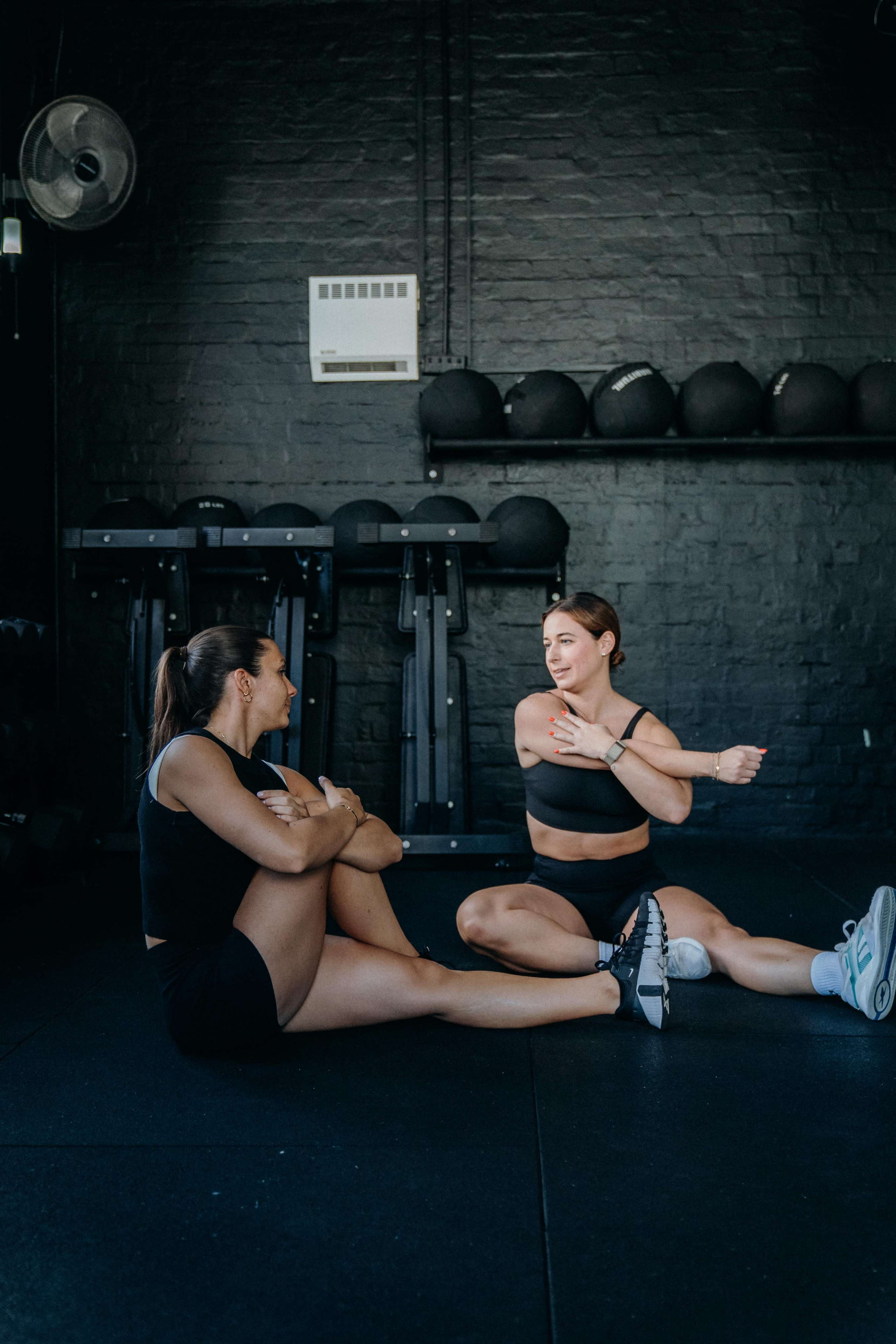 Two women sit on a gym floor in workout attire, engaging in conversation while surrounded by exercise equipment.