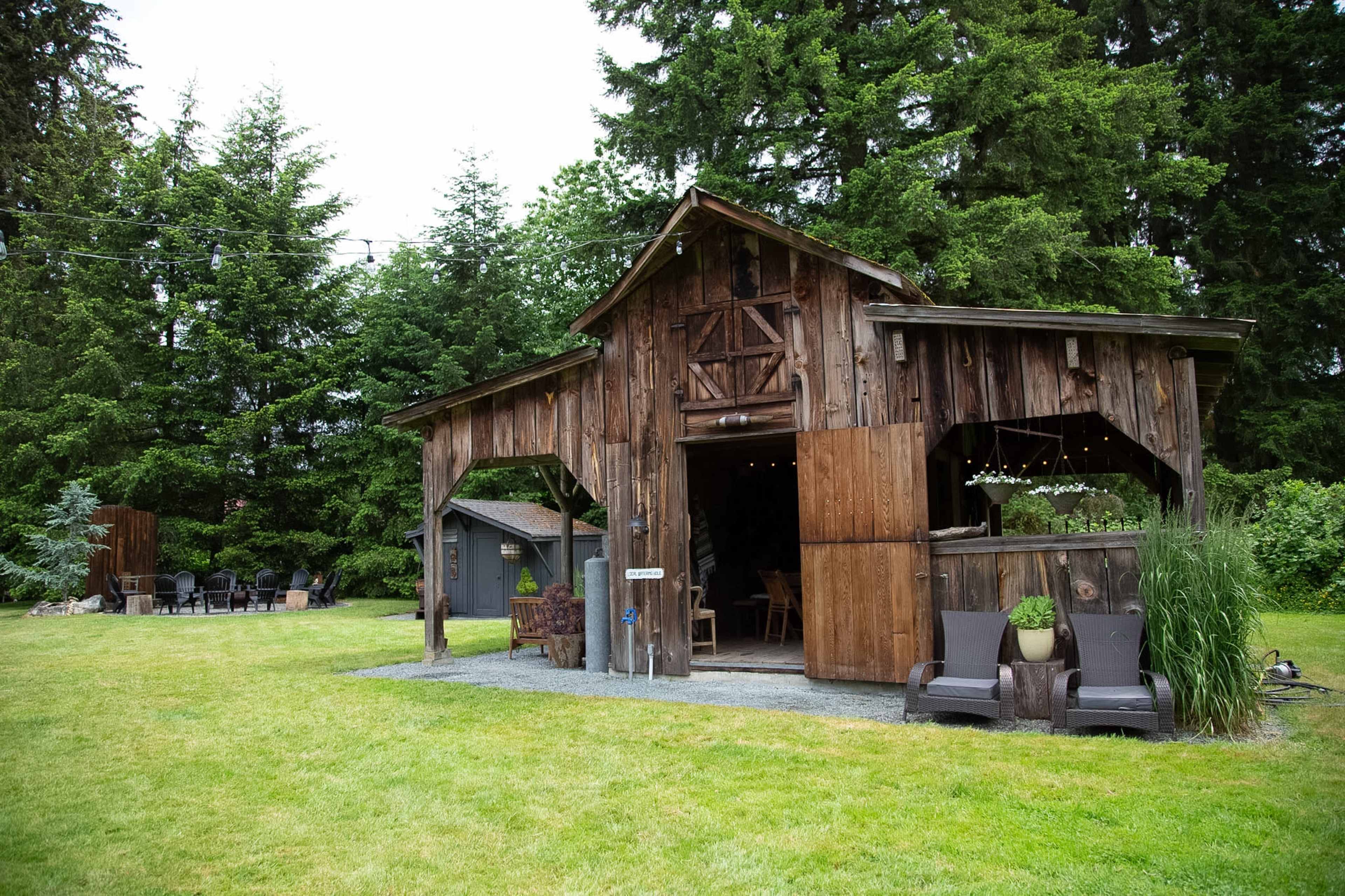 A weathered wooden barn is surrounded by lush green grass and trees, with seating arrangements visible nearby.