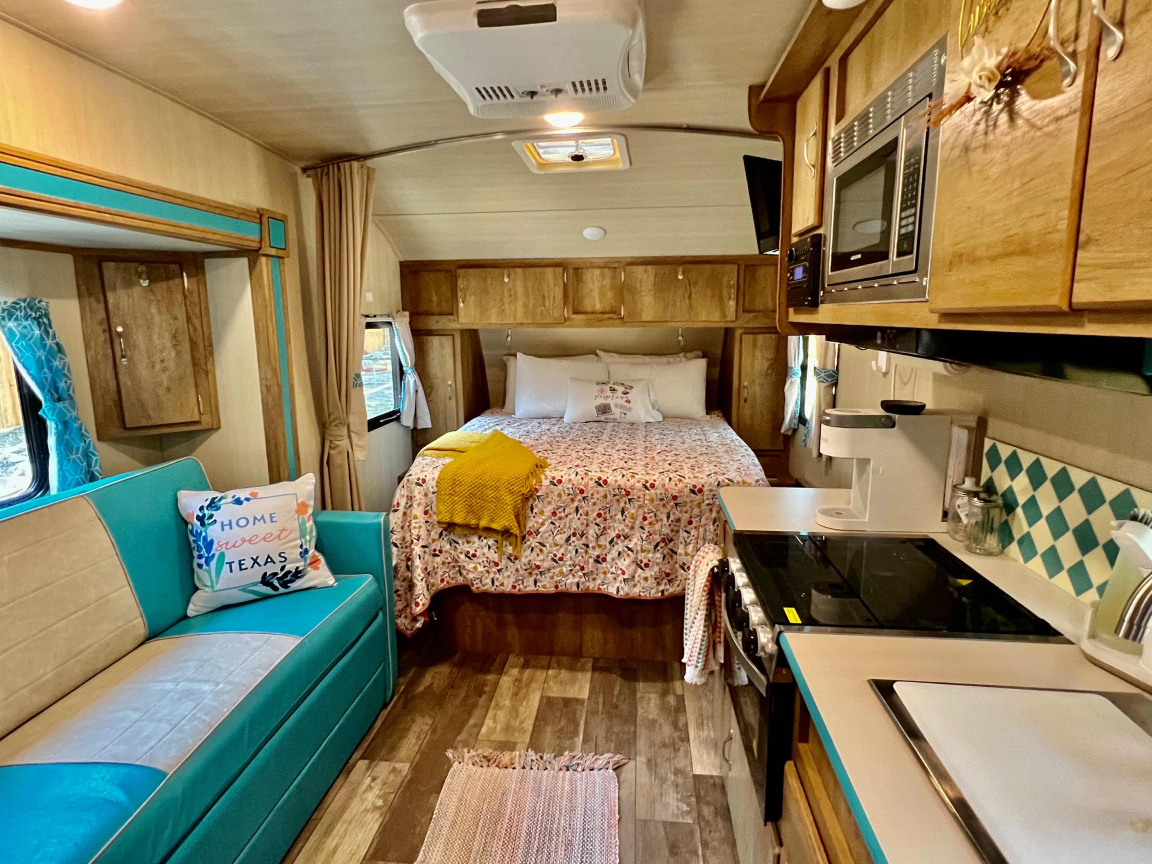 The interior of a vintage camper features a bed with floral bedding, a small kitchenette with modern appliances, and a seating area with a blue couch.