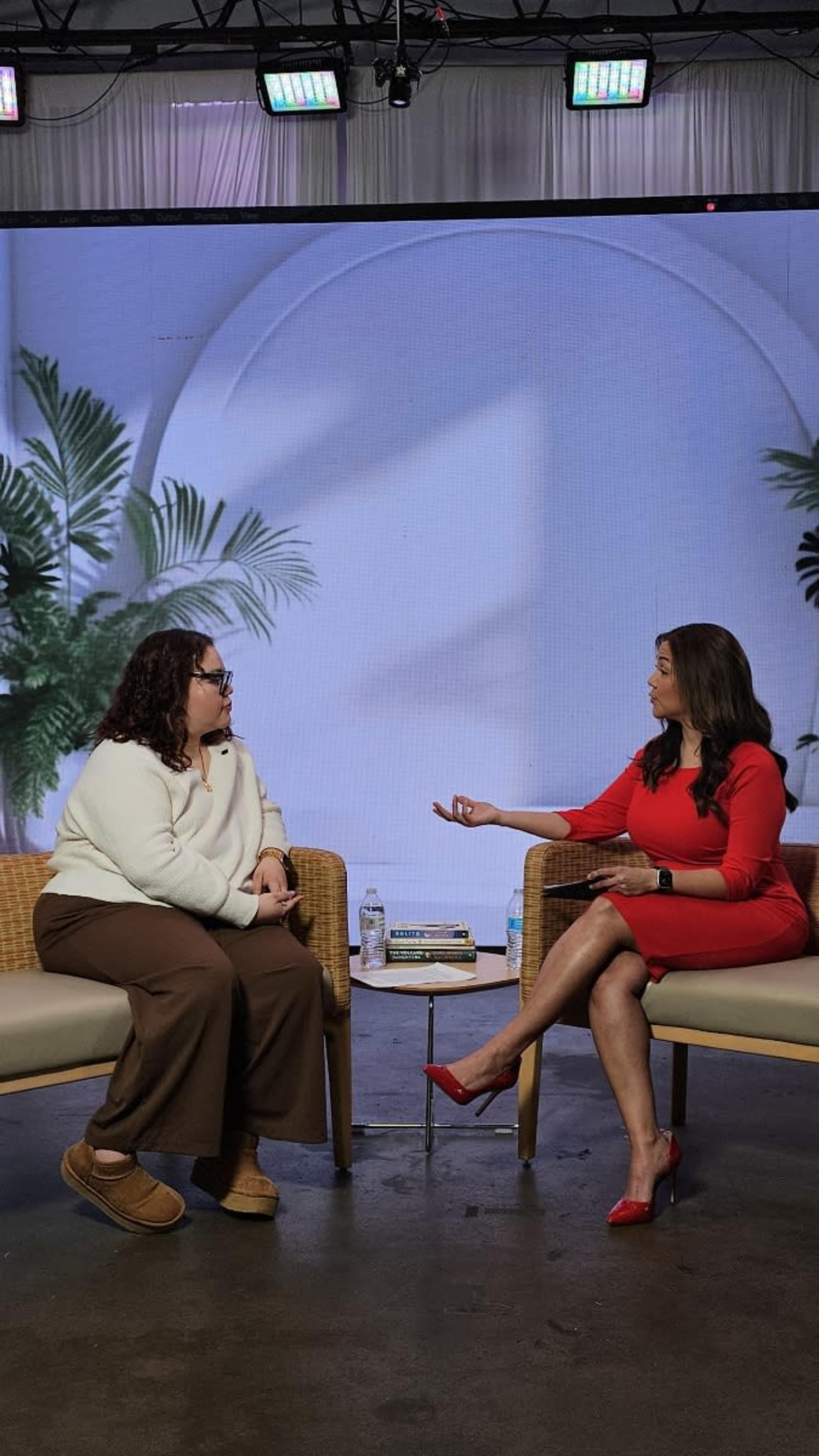 Two women are seated in a modern studio setting, one in a red dress and the other in a white sweater and brown pants, while discussing in front of a backdrop featuring plants.