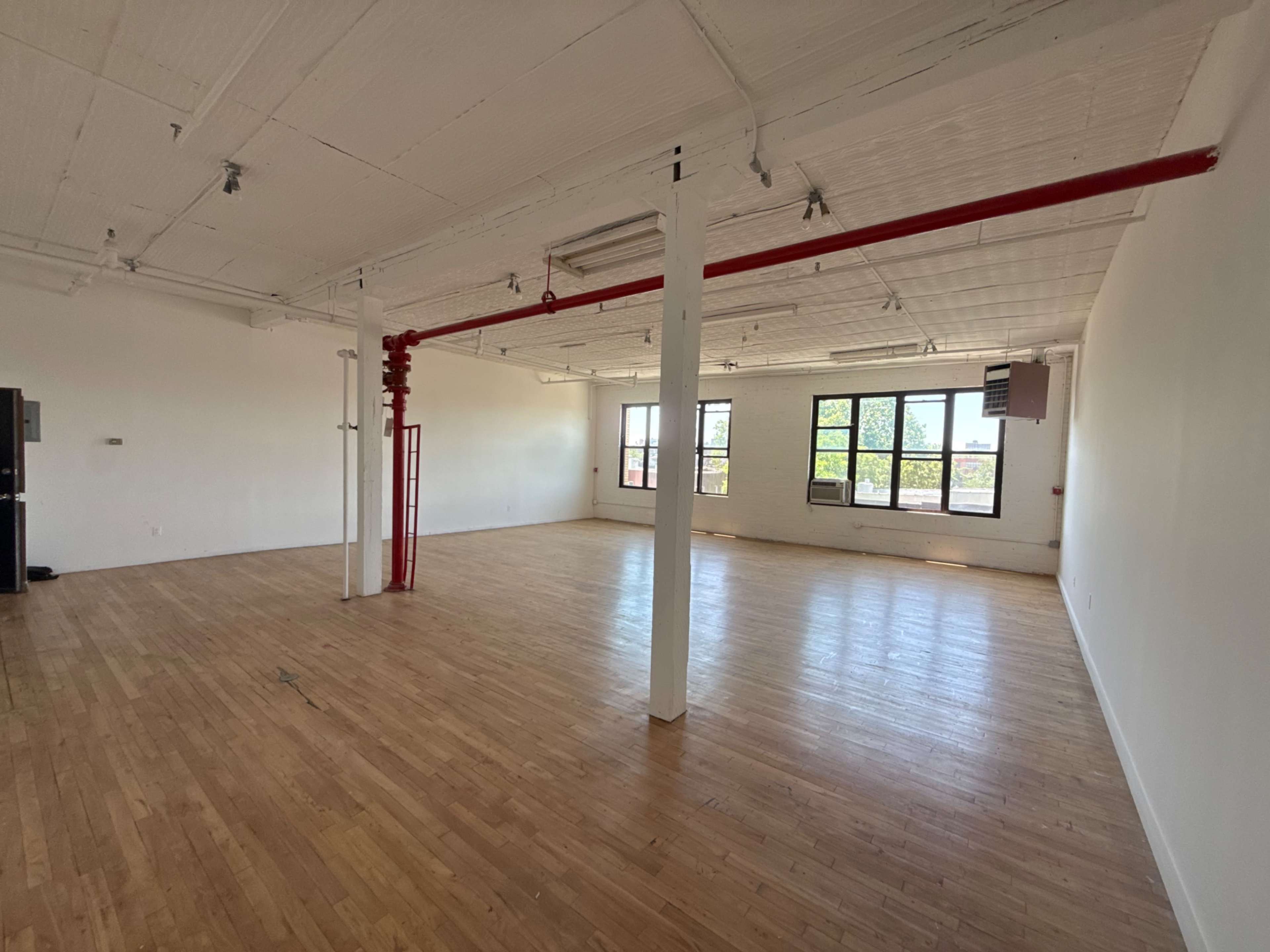 The image shows an empty, spacious room with wooden floors and large windows.