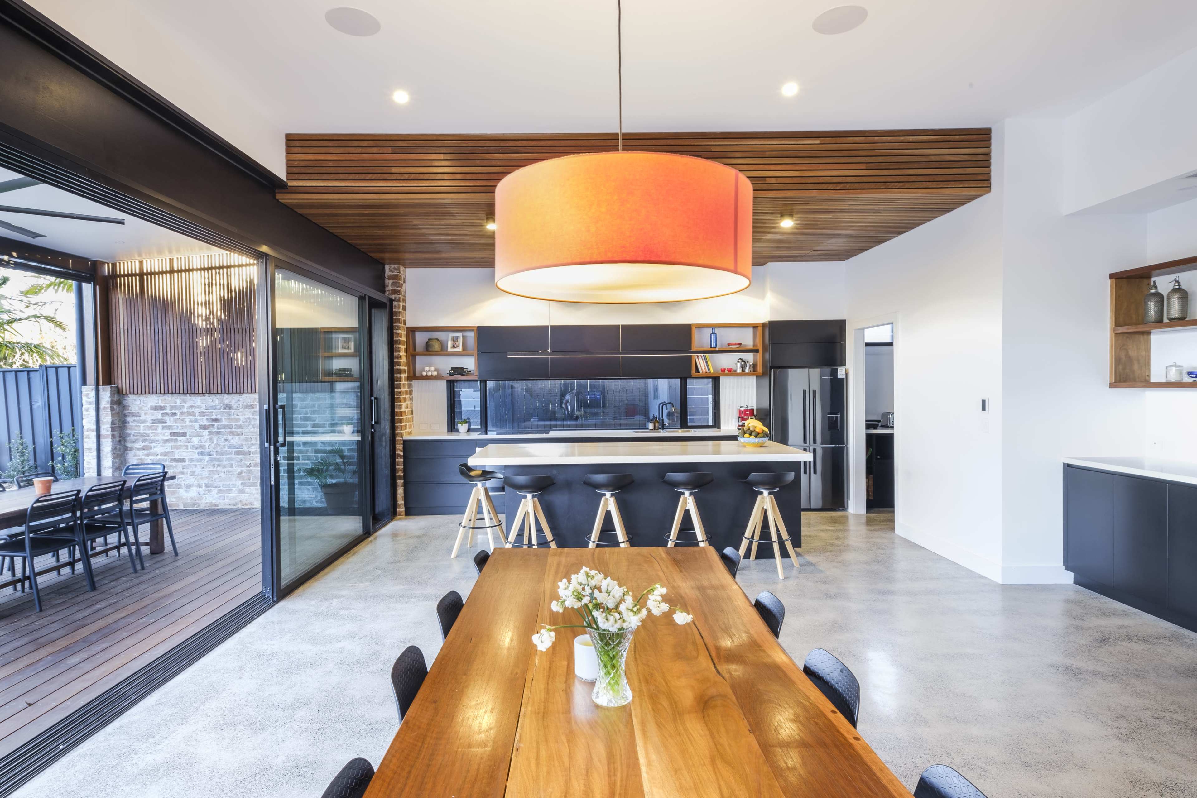 A modern kitchen and dining area featuring a large wooden table, black bar stools at a kitchen island, and large glass doors leading to an outdoor patio.