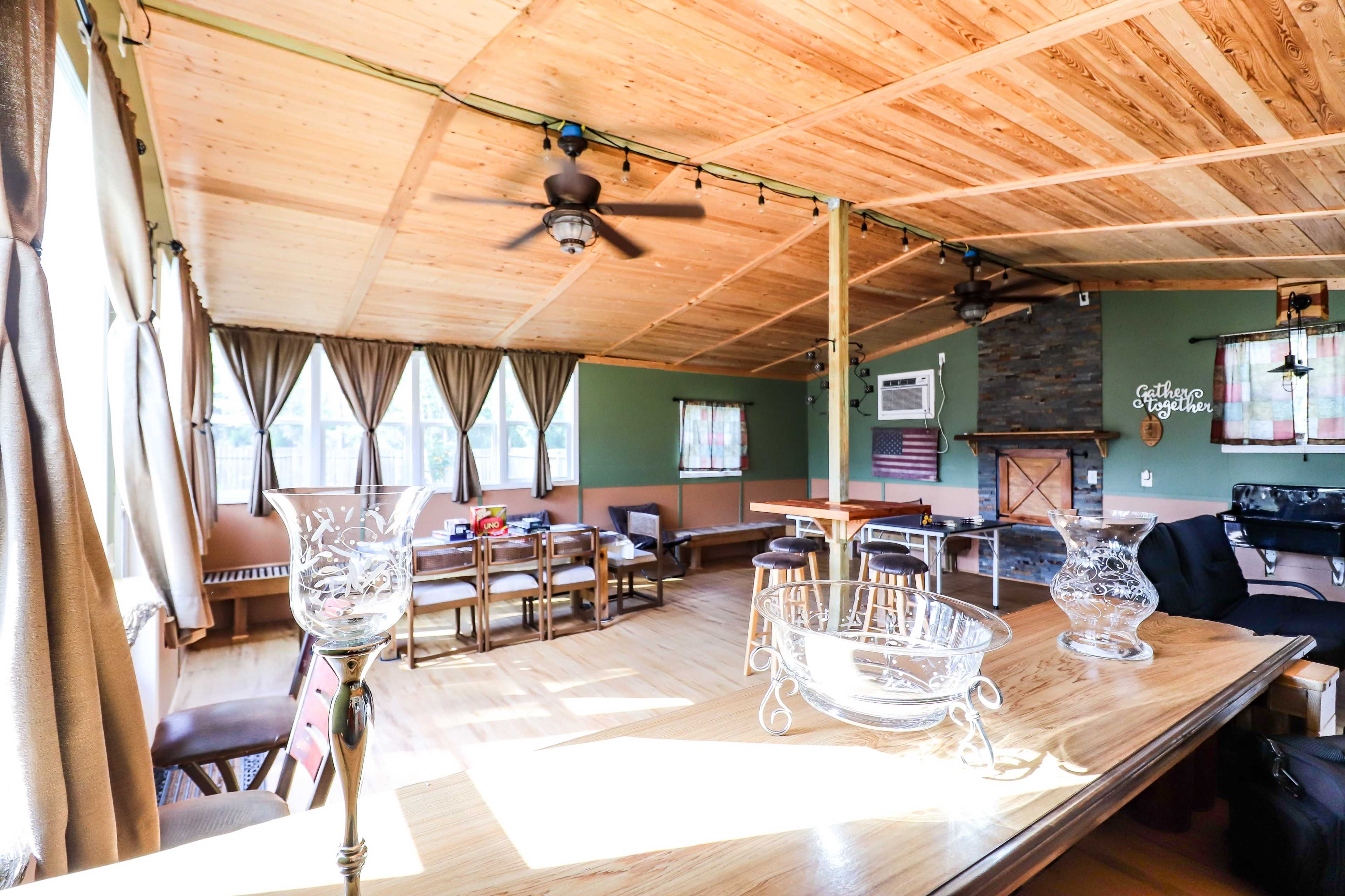 The image shows a spacious interior room with wooden ceilings, large windows, and a combination of dining and lounge furniture.