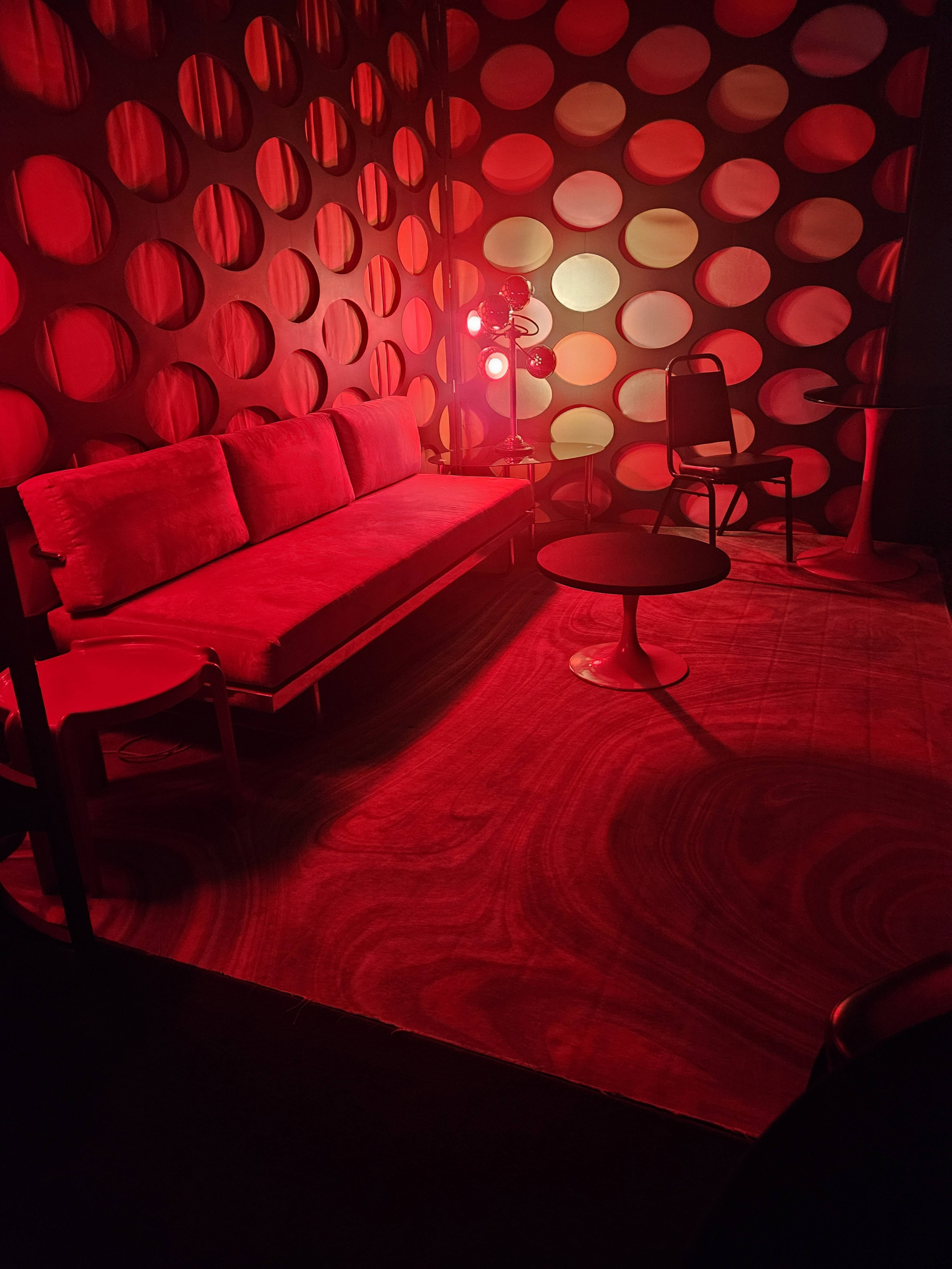 A dimly lit room features a red color scheme, with a sofa, a table, and chairs arranged against a wall adorned with circular patterns.