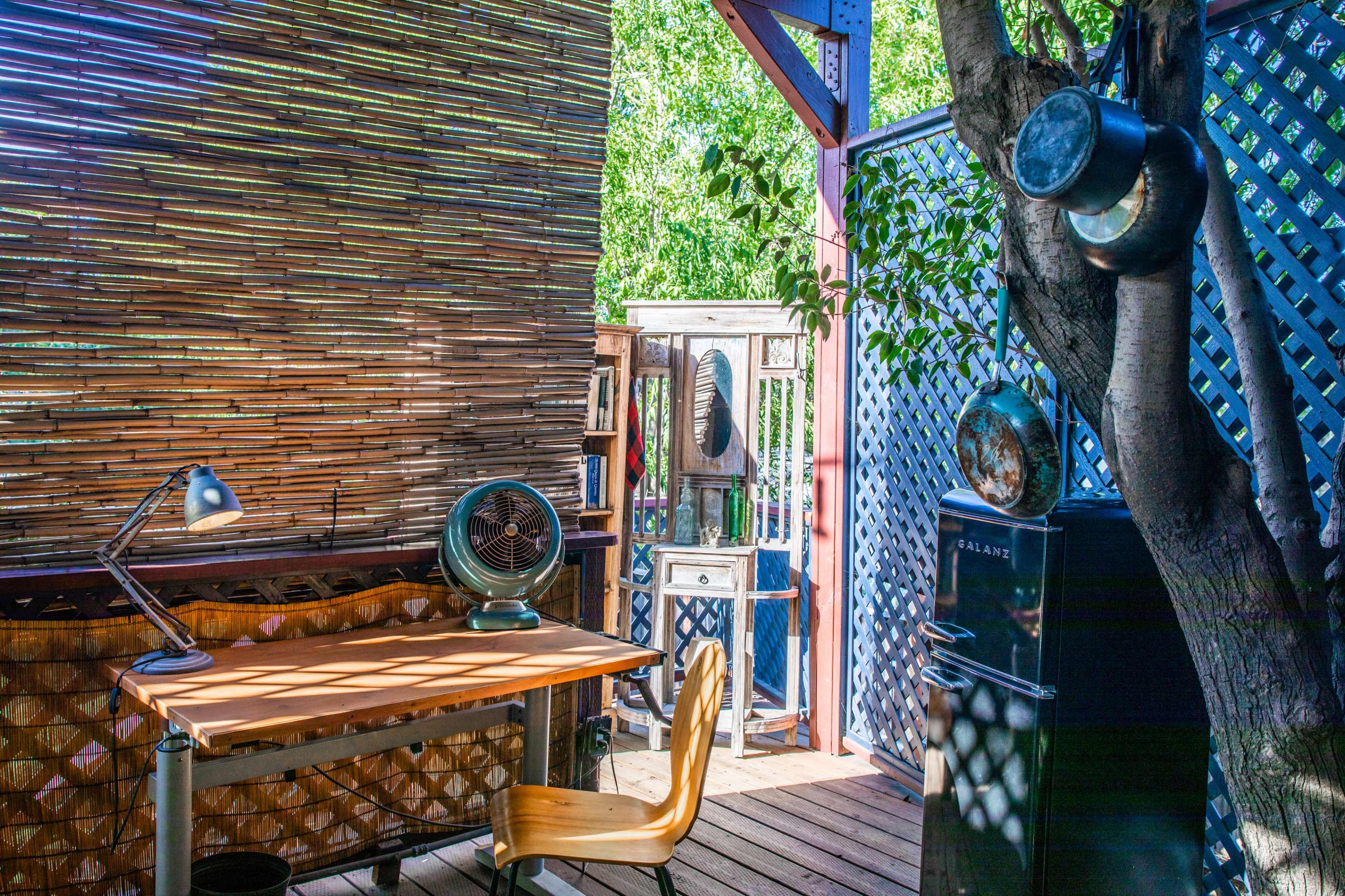 A wooden desk and chair are positioned in a bright outdoor setting with bamboo walls, flanked by a vintage fan and a small refrigerator under a tree.