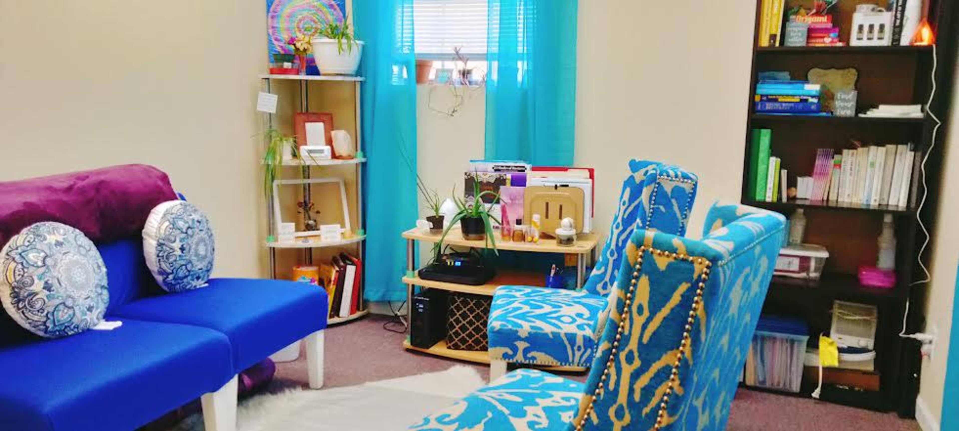 A cozy interior space with two blue chairs, a purple couch, a bookshelf filled with books, and plants arranged on a shelf.