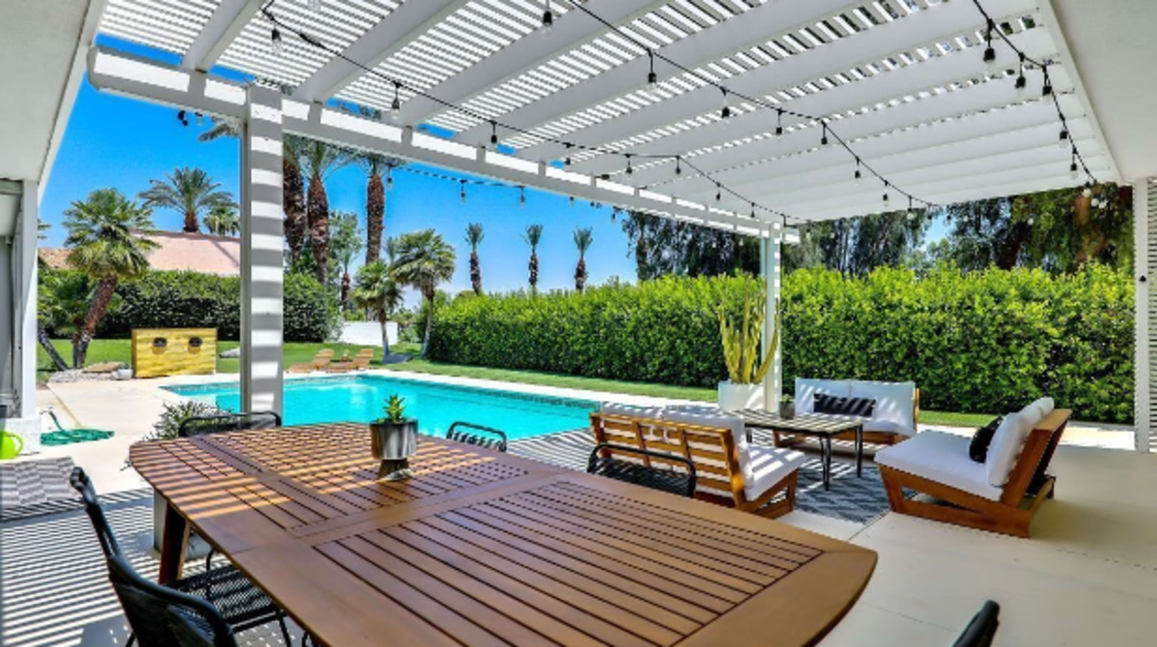 The image shows a covered patio with a wooden dining table and seating area overlooking a swimming pool and landscaped garden.