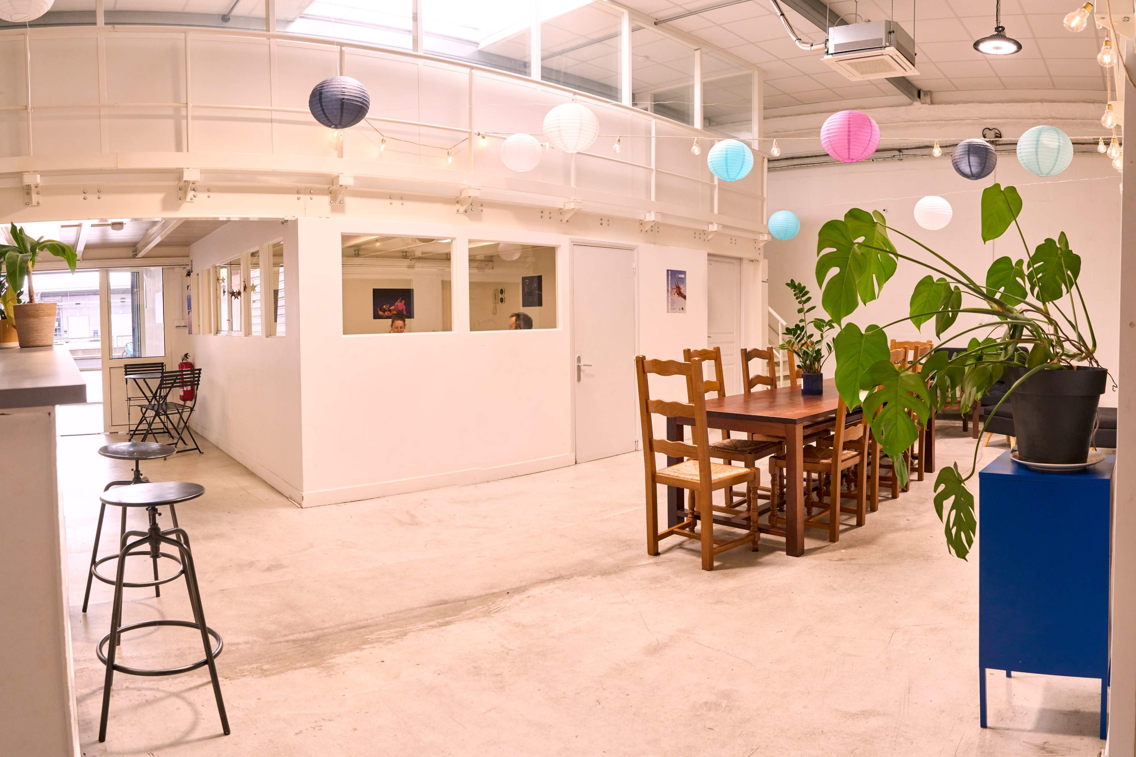 The image shows a brightly lit communal space featuring a long wooden dining table, several chairs, and decorative paper lanterns hanging from the ceiling.