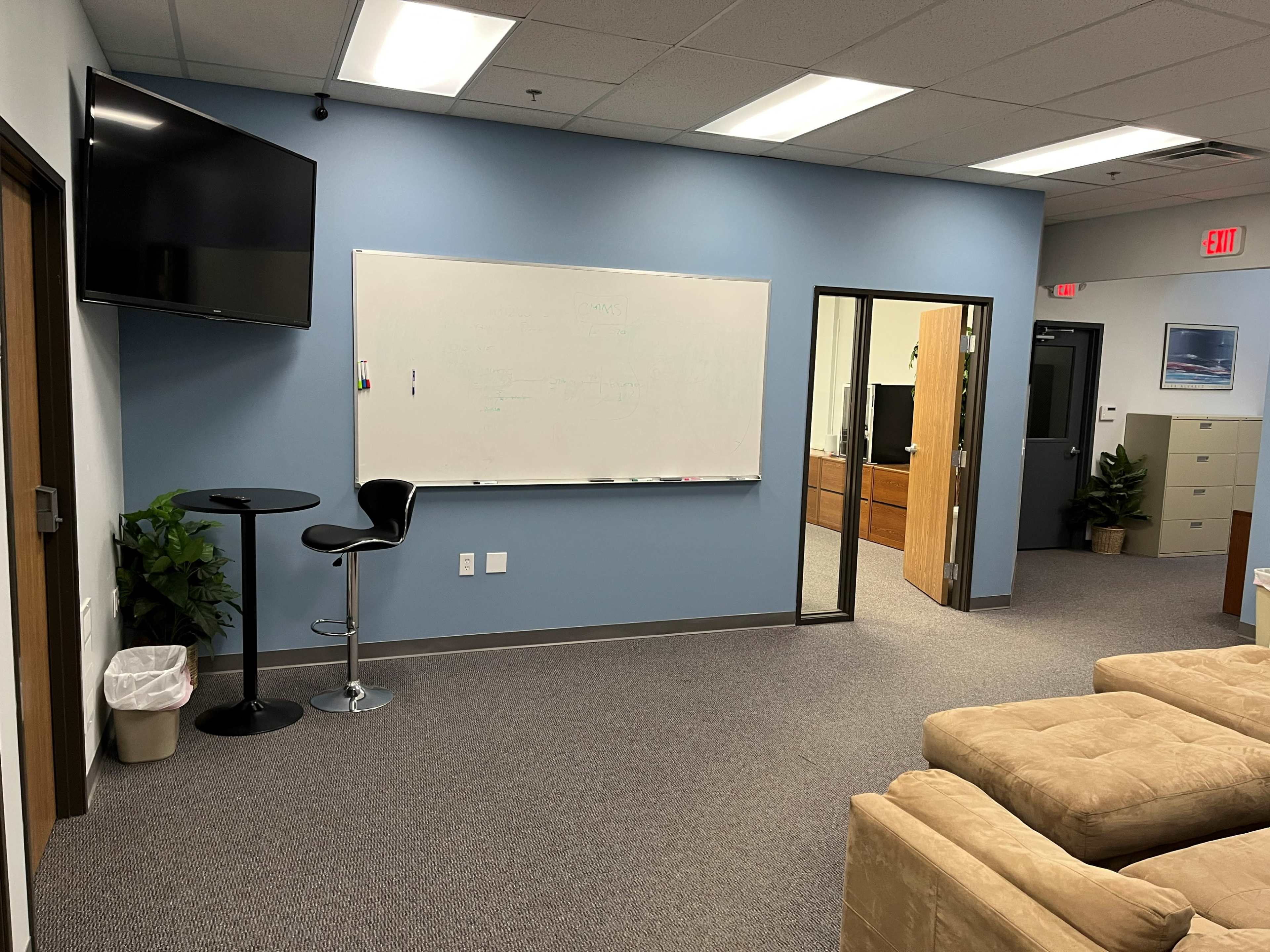 An office space with a seating area, a whiteboard, a bar table with a stool, a television mounted on the wall, and a doorway leading to another room.