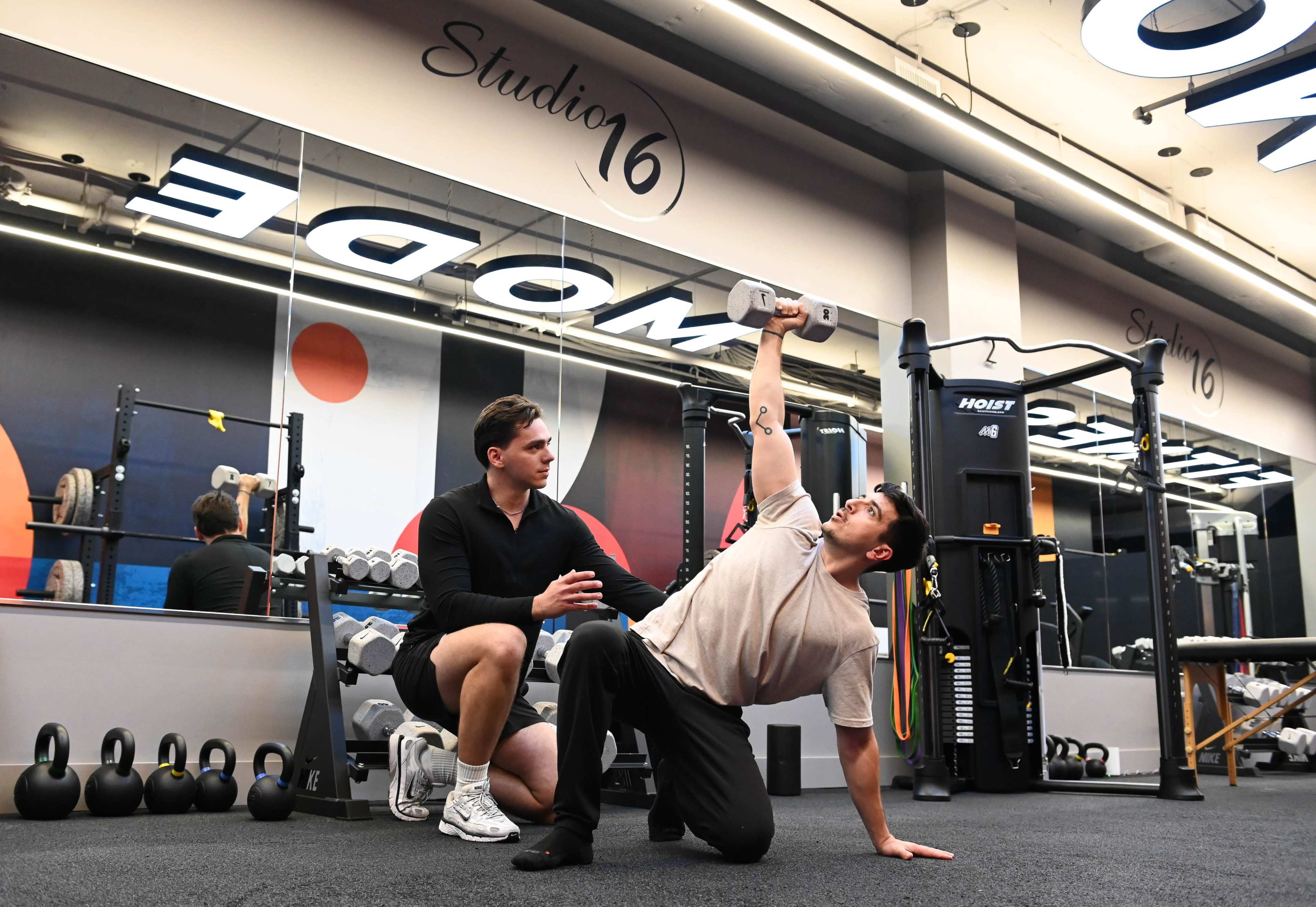 A personal trainer assists a client performing a single-arm dumbbell press in a fitness studio with gym equipment and mirrors.