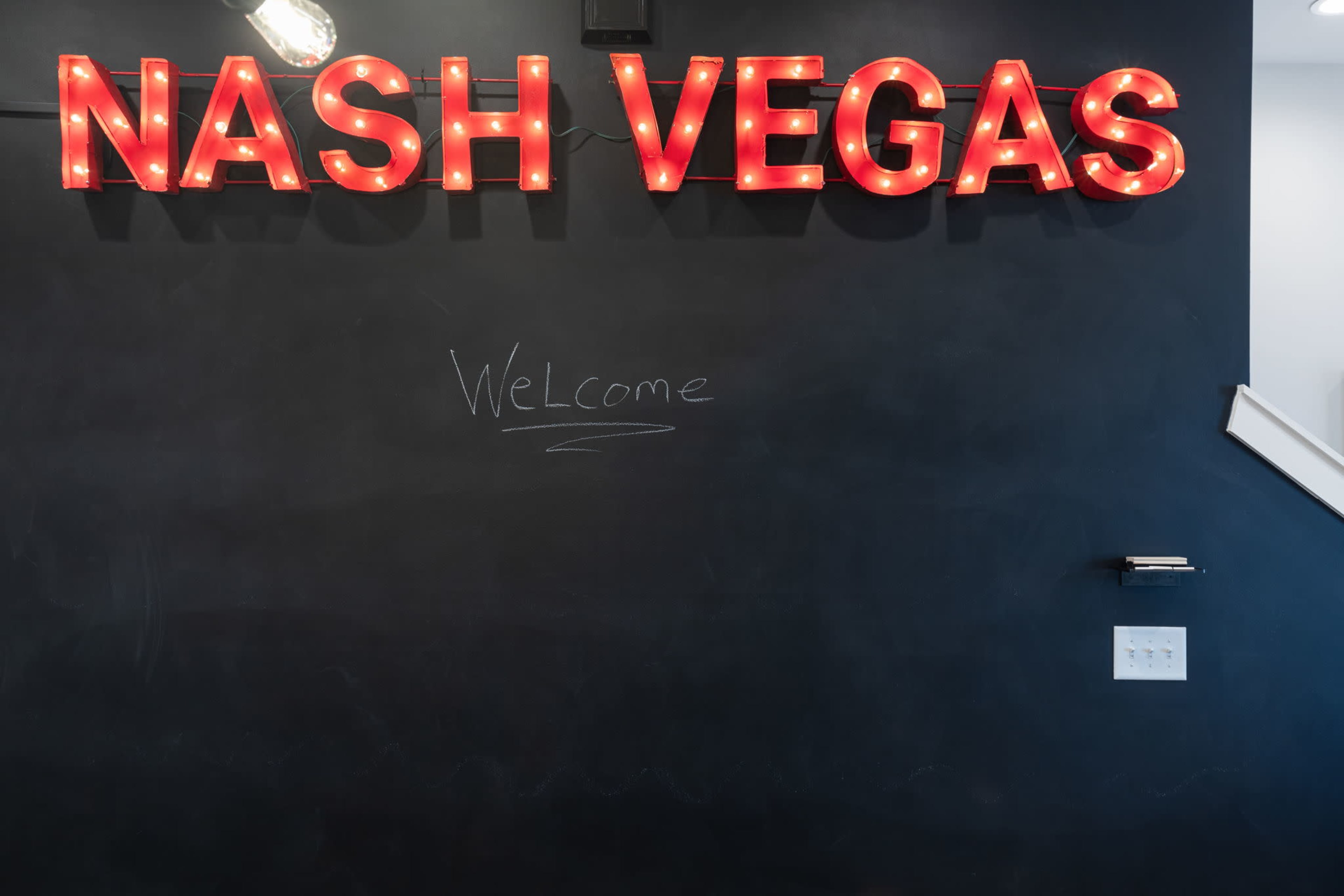 A wall displays the illuminated sign "NASH VEGAS" in red letters above a chalkboard with the word "Welcome" written in white chalk.