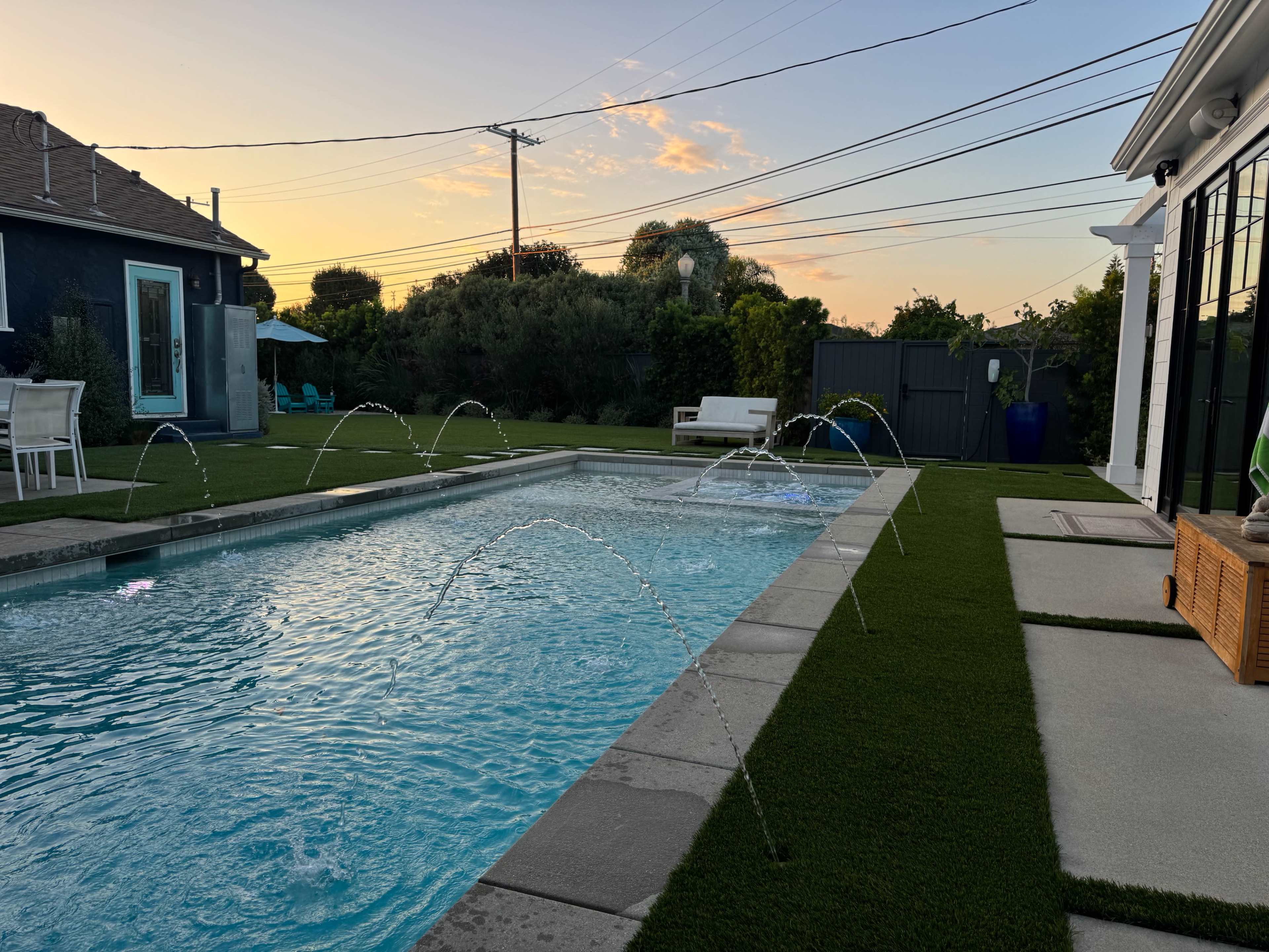 The image shows a swimming pool with water fountains set against a backdrop of a sunset, surrounded by artificial grass and seating areas.