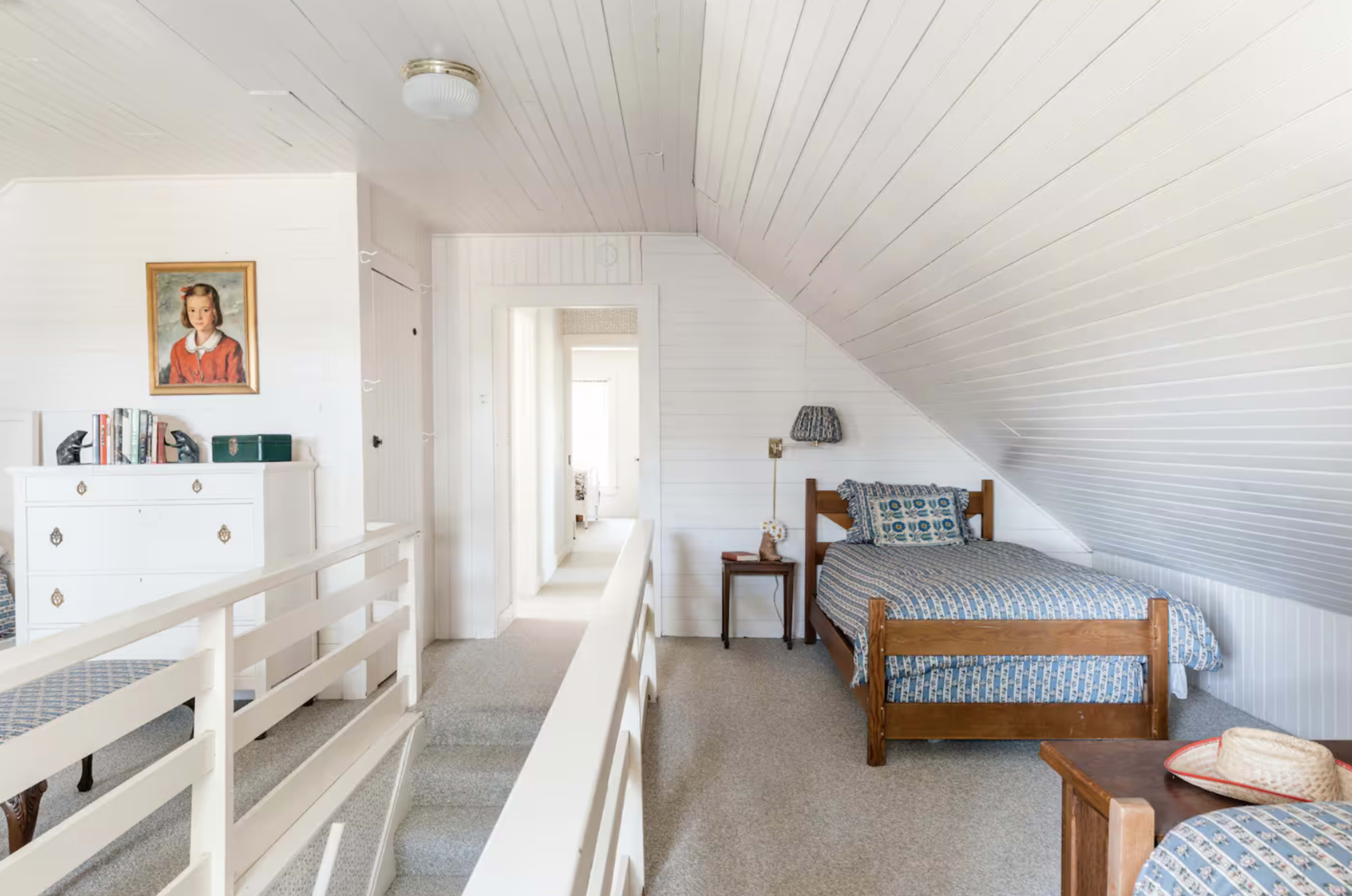The image shows a bright, white attic bedroom featuring a bed, a small side table, and a dresser, with sloped ceilings and a portrait on the wall.
