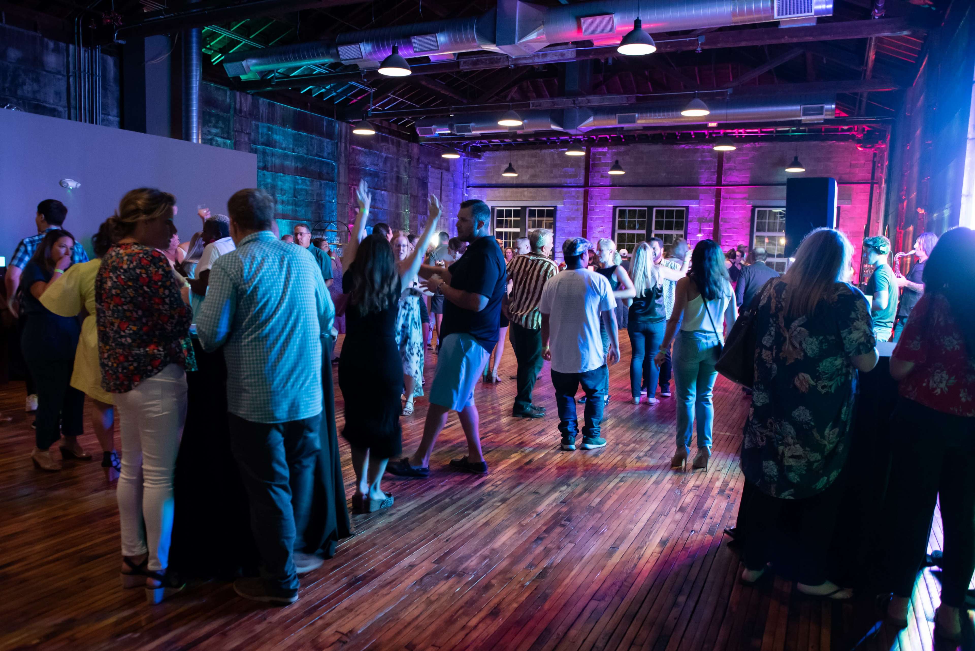 A large group of people mingles at a social event inside a well-lit venue with wooden floors and colorful lighting.