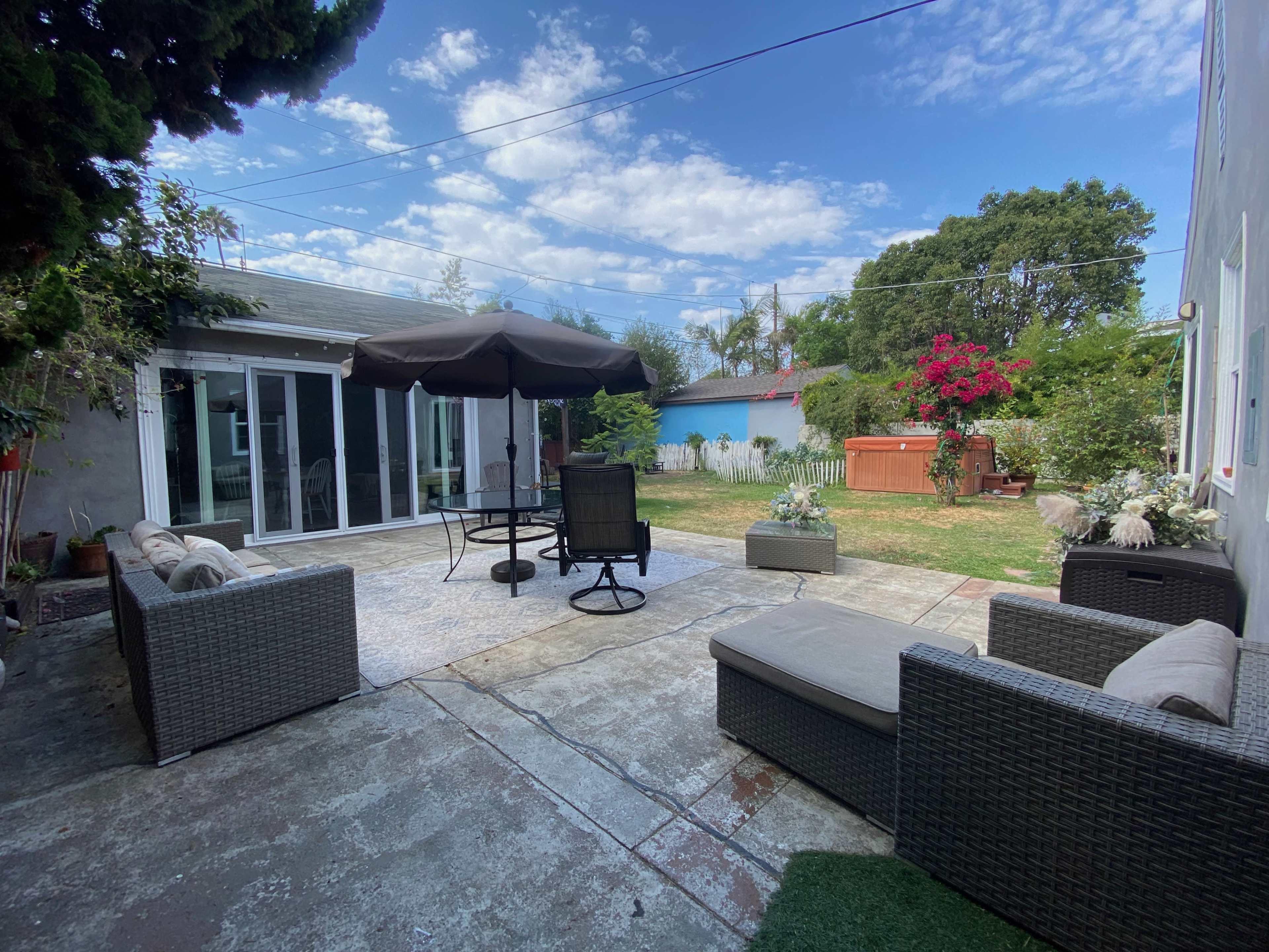 A patio area furnished with seating, a table with an umbrella, and a lawn surrounded by greenery and flowering plants.