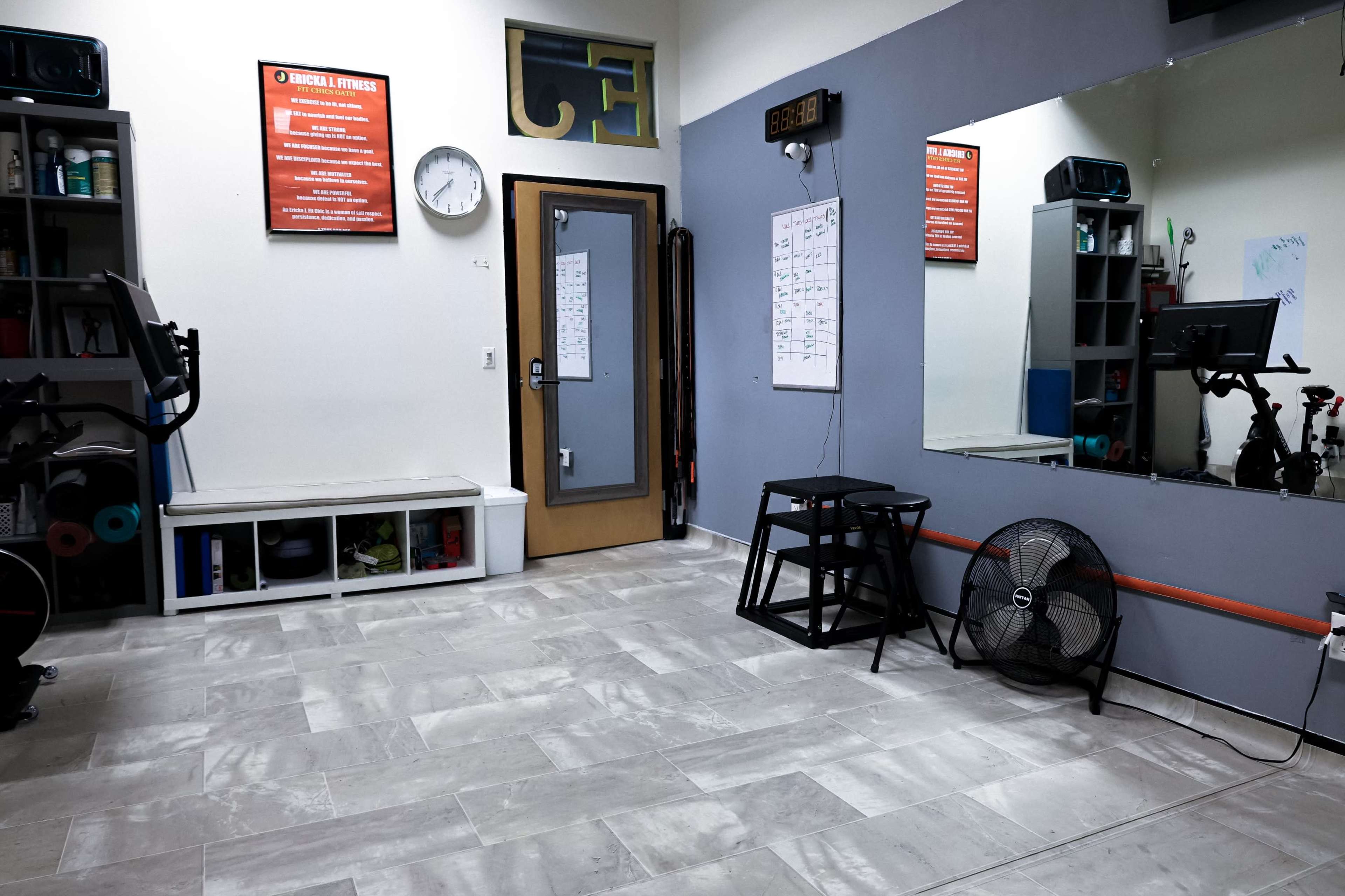 A small fitness room equipped with exercise equipment, a clock, a whiteboard, and storage units along the walls.
